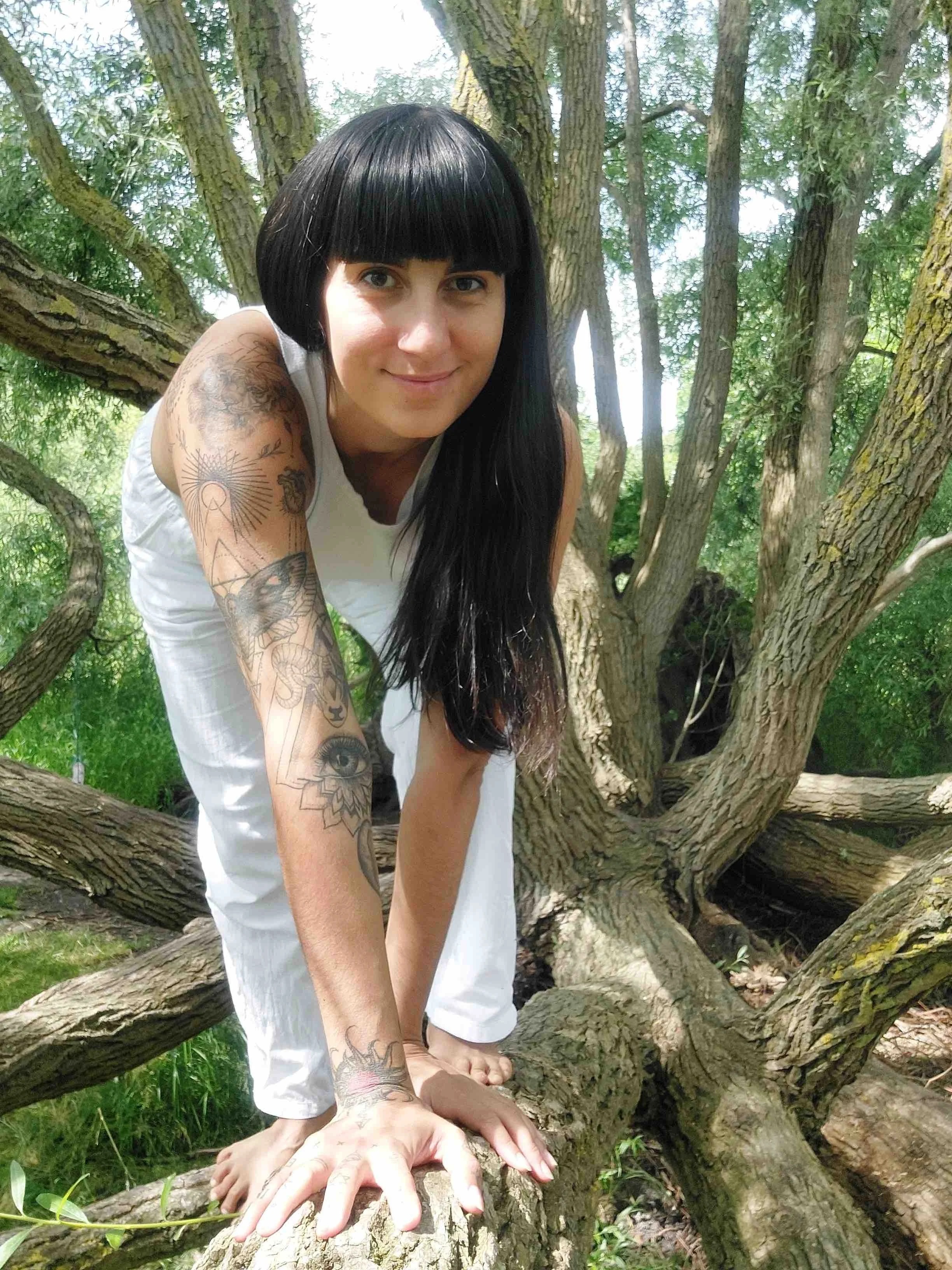 A woman with long black hair and tattoos on her arm, smiling, standing in a tree with multiple branches and green foliage in the background.