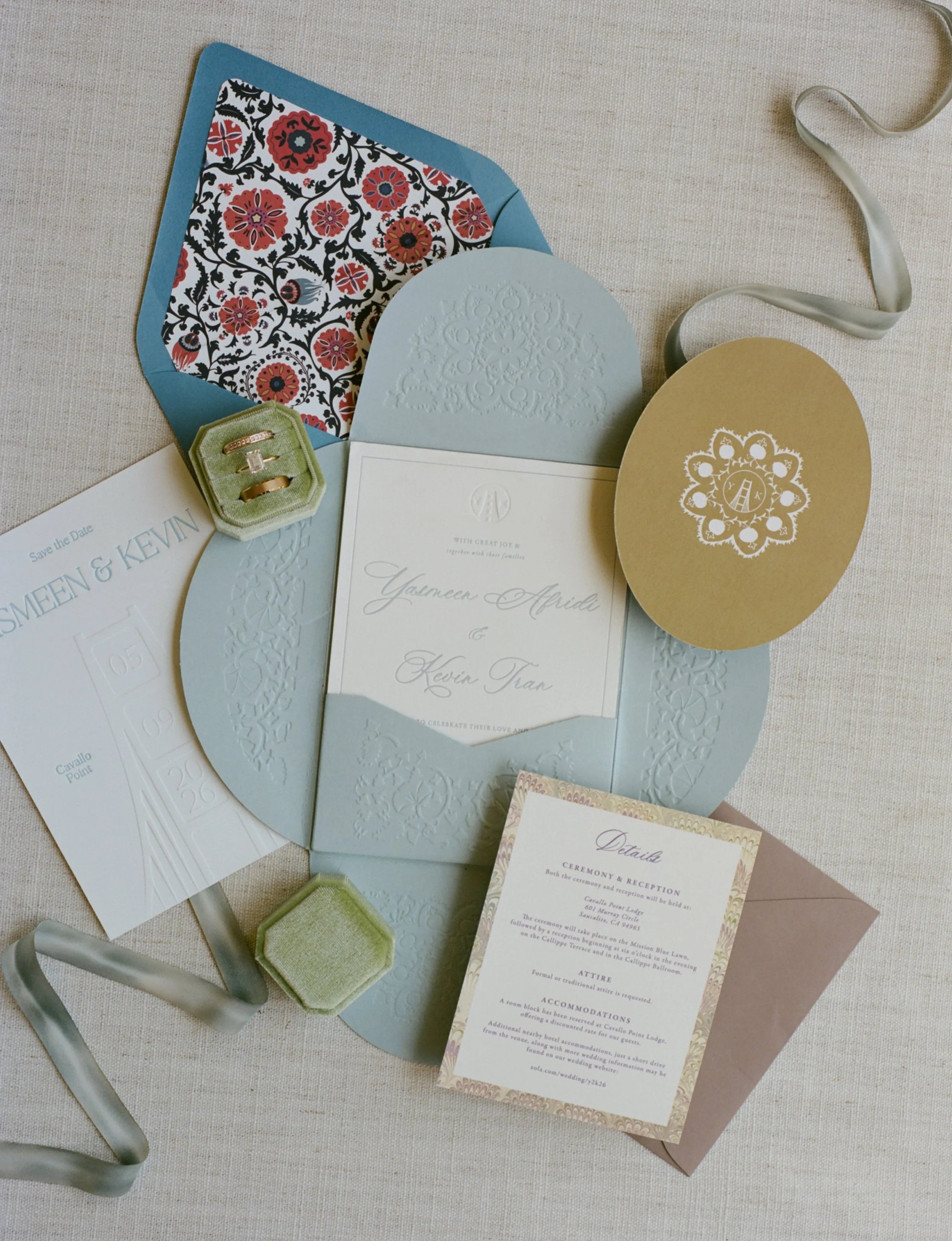 Wedding invitation stationery with a floral motif, rings in a green velvet box, a ribbon, and a detailed ceremony and reception information card, all placed on a light-colored fabric surface.
