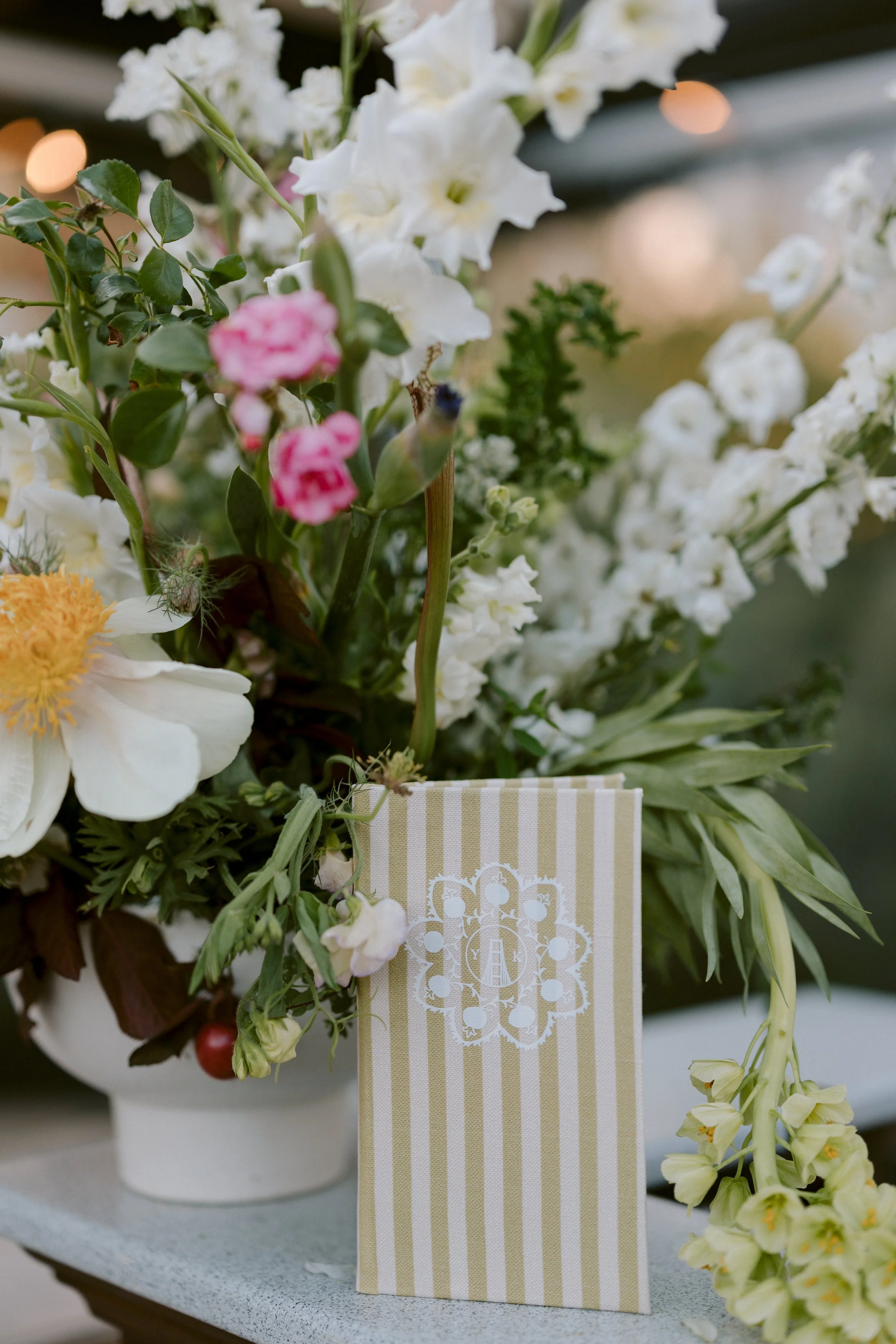Flower arrangement with white, pink, yellow, and green flowers in a white pot, with a decorative card hanging from the arrangement.