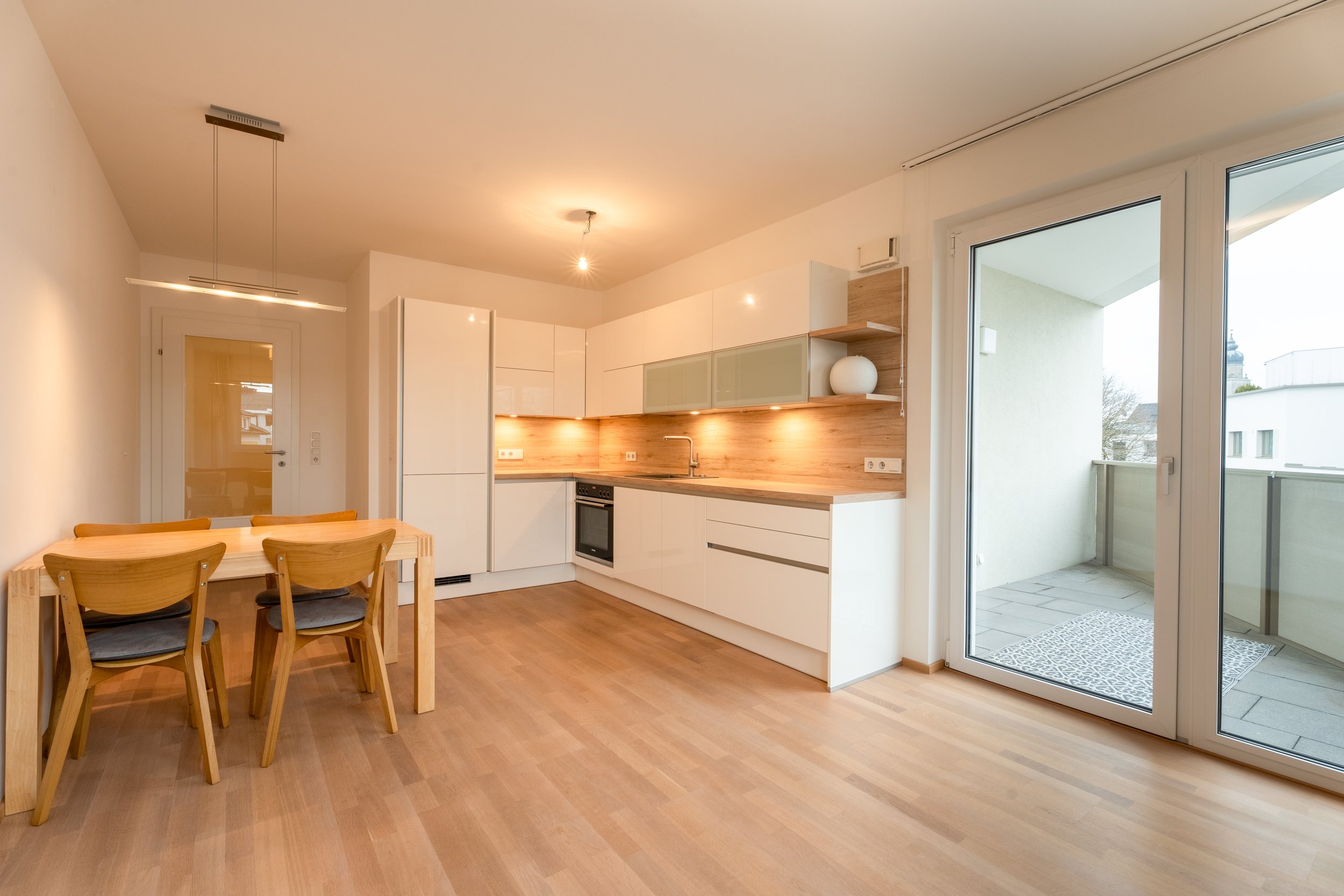 Moderne Küche und Essbereich mit Holzfußboden, weißen Schränken, Holzwand und Balkon.