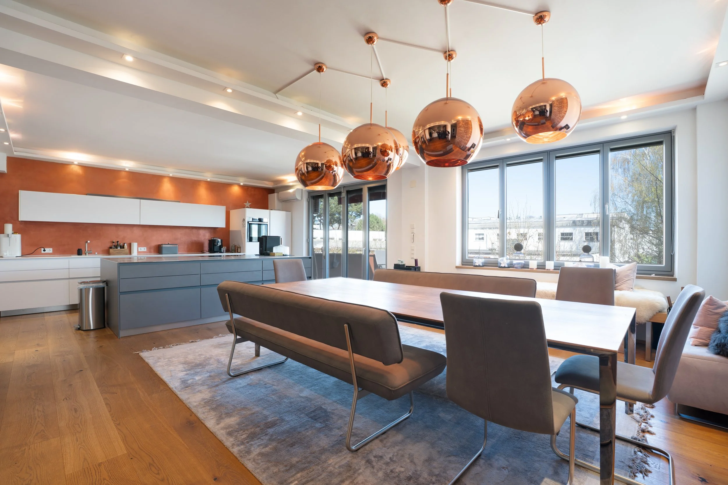 Moderner Ess- und Küchenbereich mit Holztisch, bunten Sitzbänken und Stühlen, großen Fenstern, vier kupferfarbenen Hängeleuchten und einer Küchenzeile mit weißen und grauen Schränken.