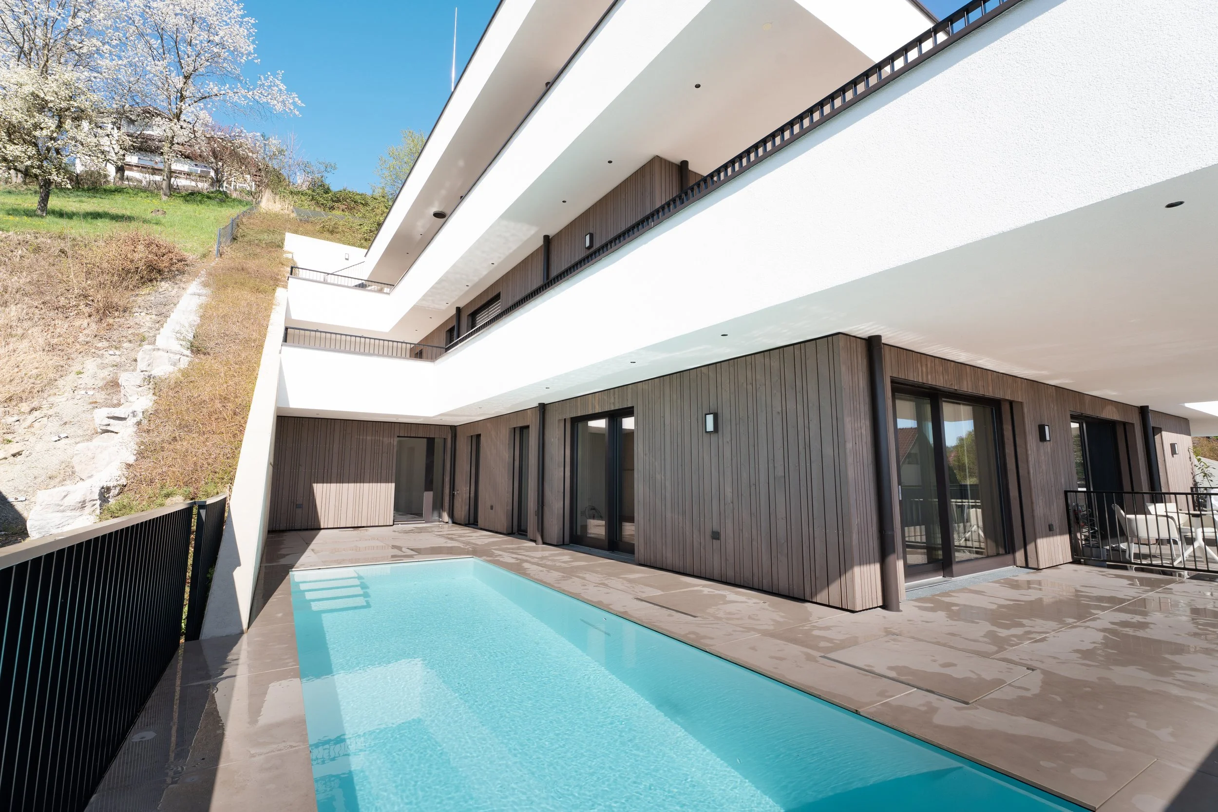 Moderne Wohnung mit Balkon, Terrasse und Swimmingpool, helles Holz und weiße Wände, im Grünen, sonniges Wetter.