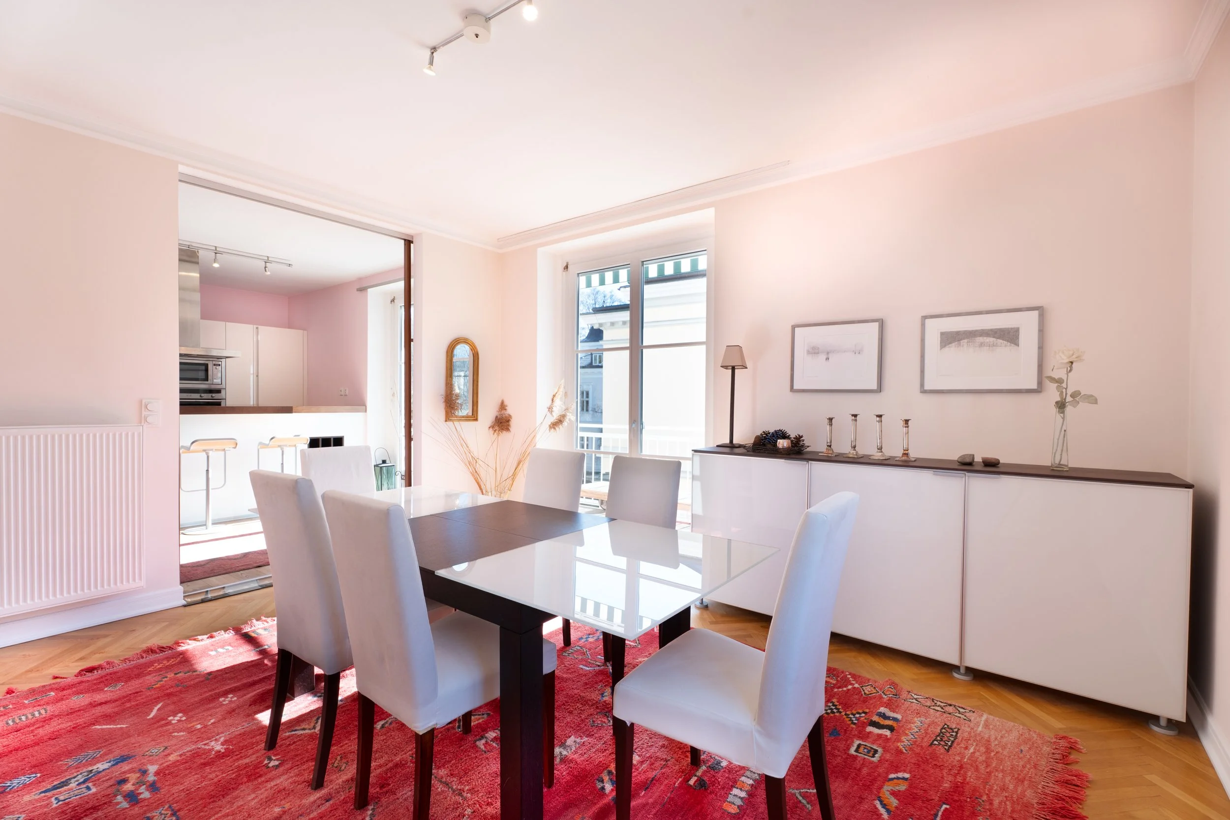 Helles Esszimmer mit weißem Tisch, sechs weißen Stühlen, rotem Teppich, modernem Sideboard mit Dekorationen, großem Fenster, Kamin und offener Küche im Hintergrund.