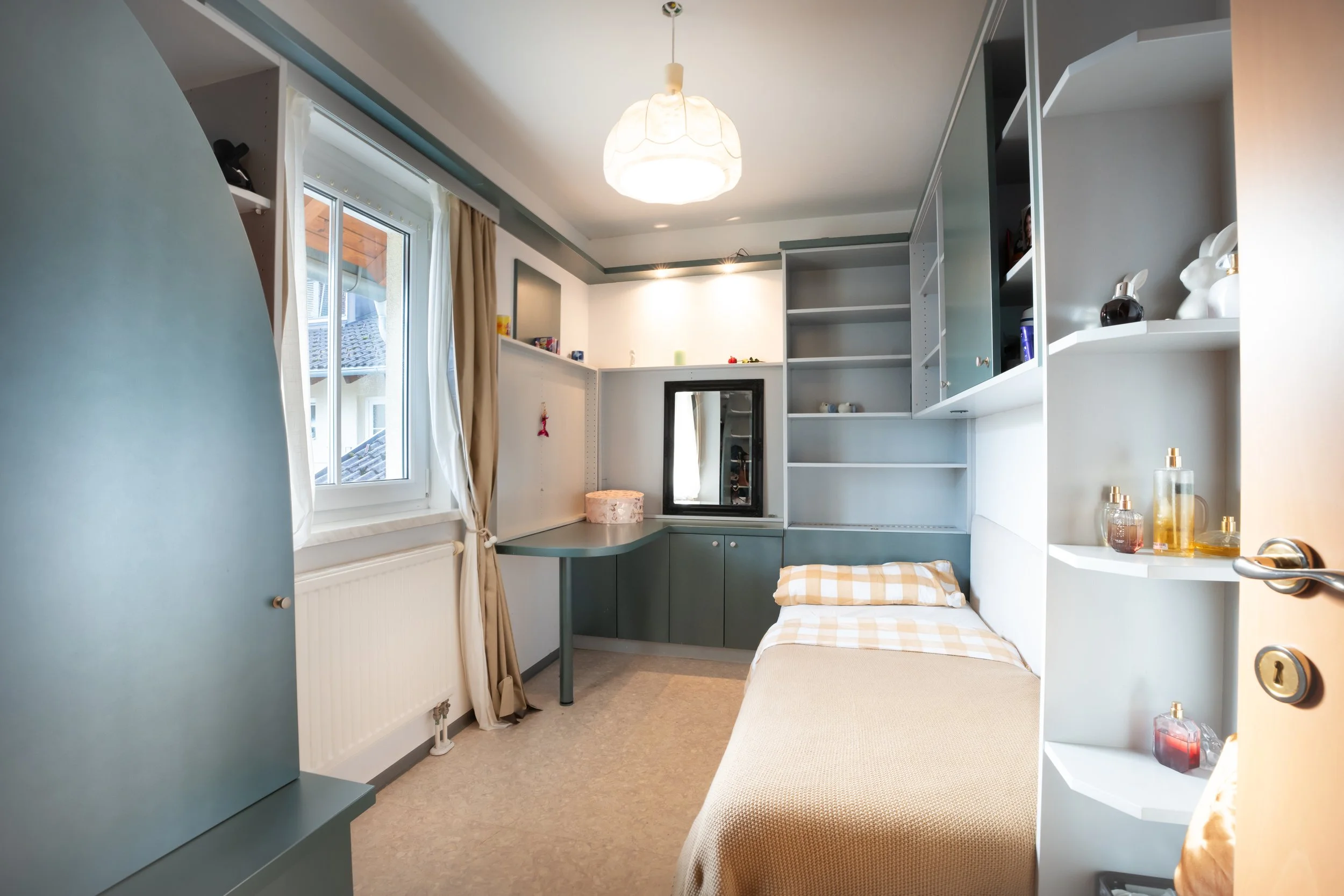 Kleines Schlafzimmer mit einem Bett, einem Fenster mit weißen Vorhängen, einem Wandspiegel, offenen Regalen und einem Schreibtisch in hellen Farben.