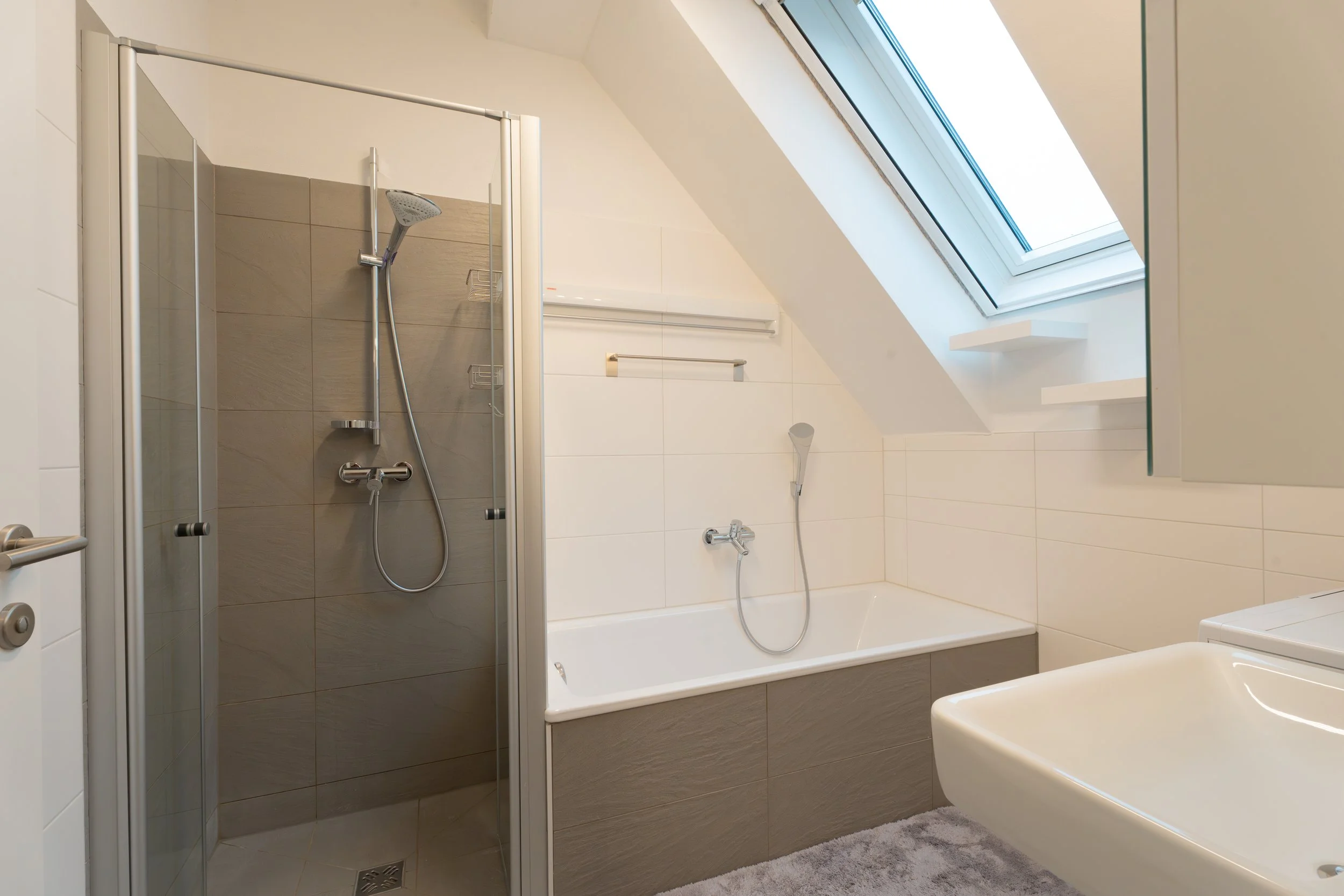 Ein modernes Badezimmer mit einer Duschecke, Badewanne, Waschbecken, Dachfenster und weißen sowie grauen Wandfliesen.