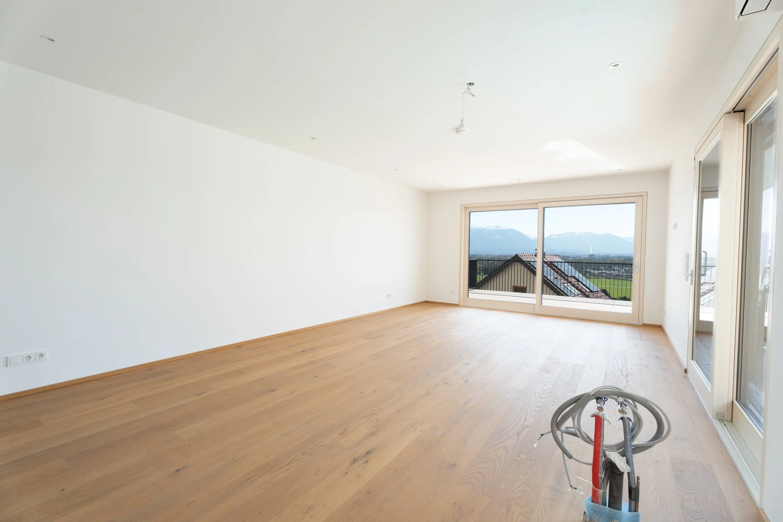 Leeres, helles Wohnzimmer mit großen Fenstern und Holzboden, Blick auf Berge und Dachkonstruktionen, Kabel im Vordergrund.