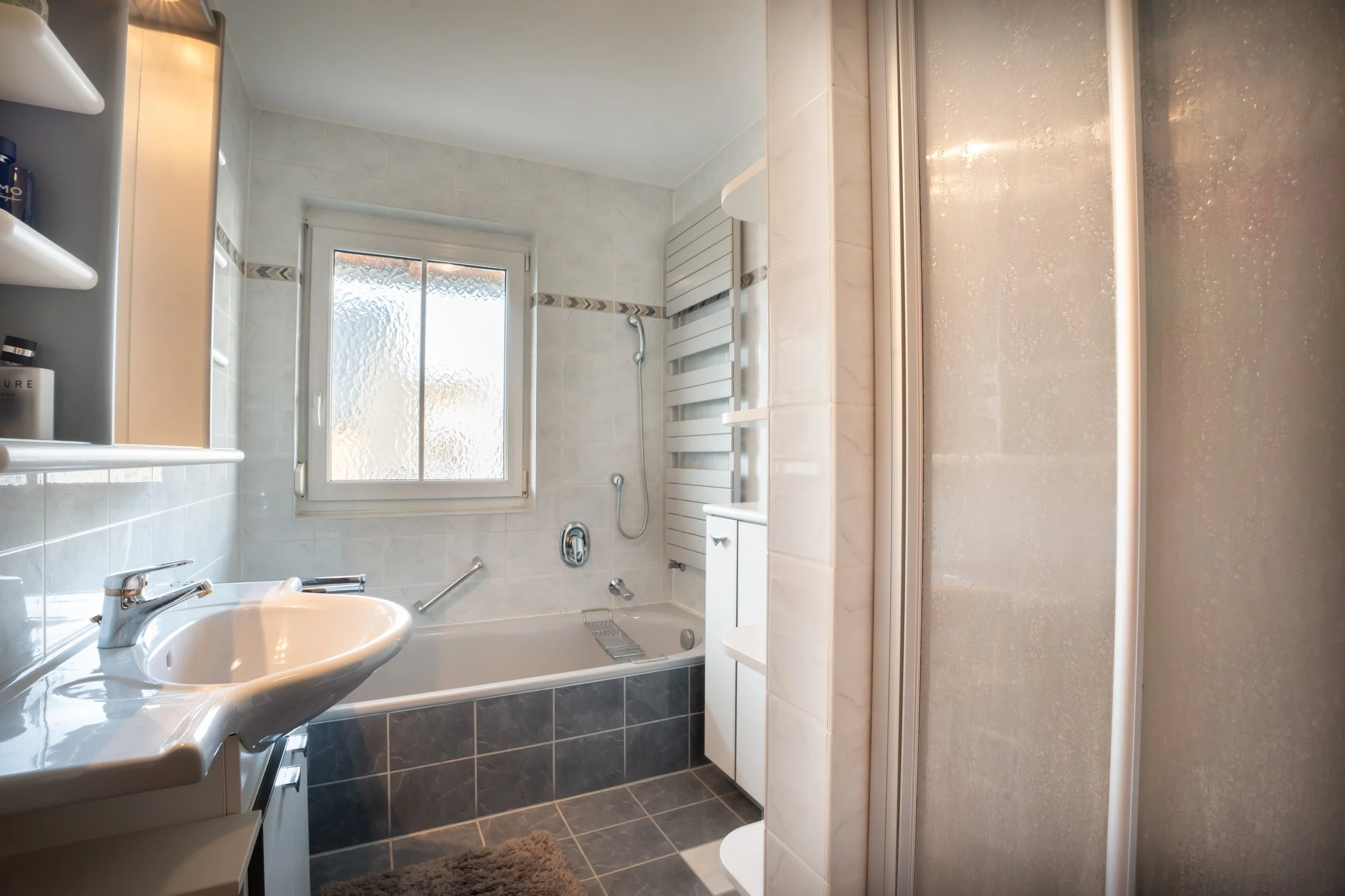 Badezimmer mit Dusche, Badewanne, Waschbecken, Fenster, Heizkörper, Regale und Handtuch, Fliesen und Beleuchtung.