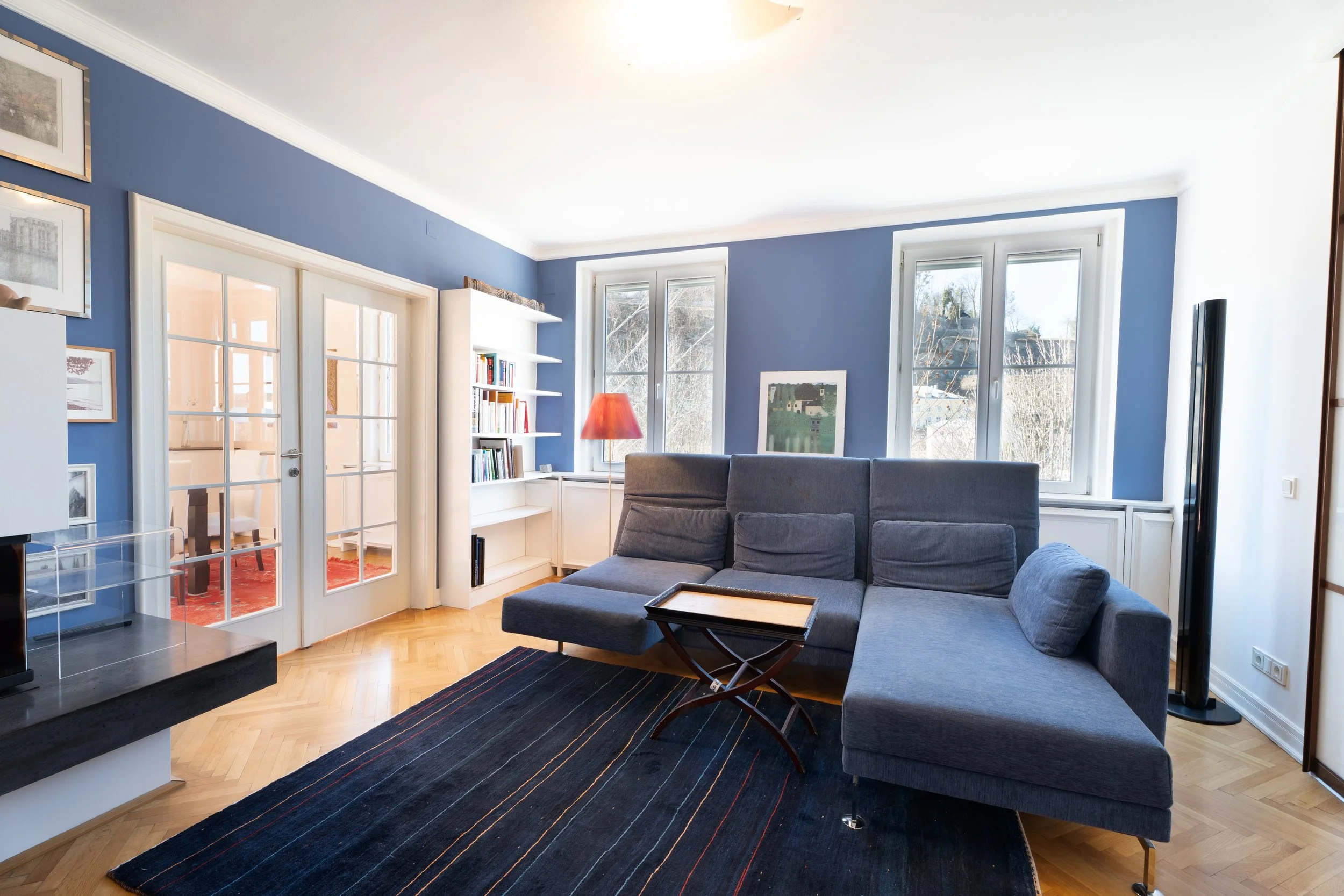 Wohnzimmer mit blauen Wänden, grauem Sofa, Fenster, Teppich, Bücherregal, Stehlampe, Dekorationsbilder.