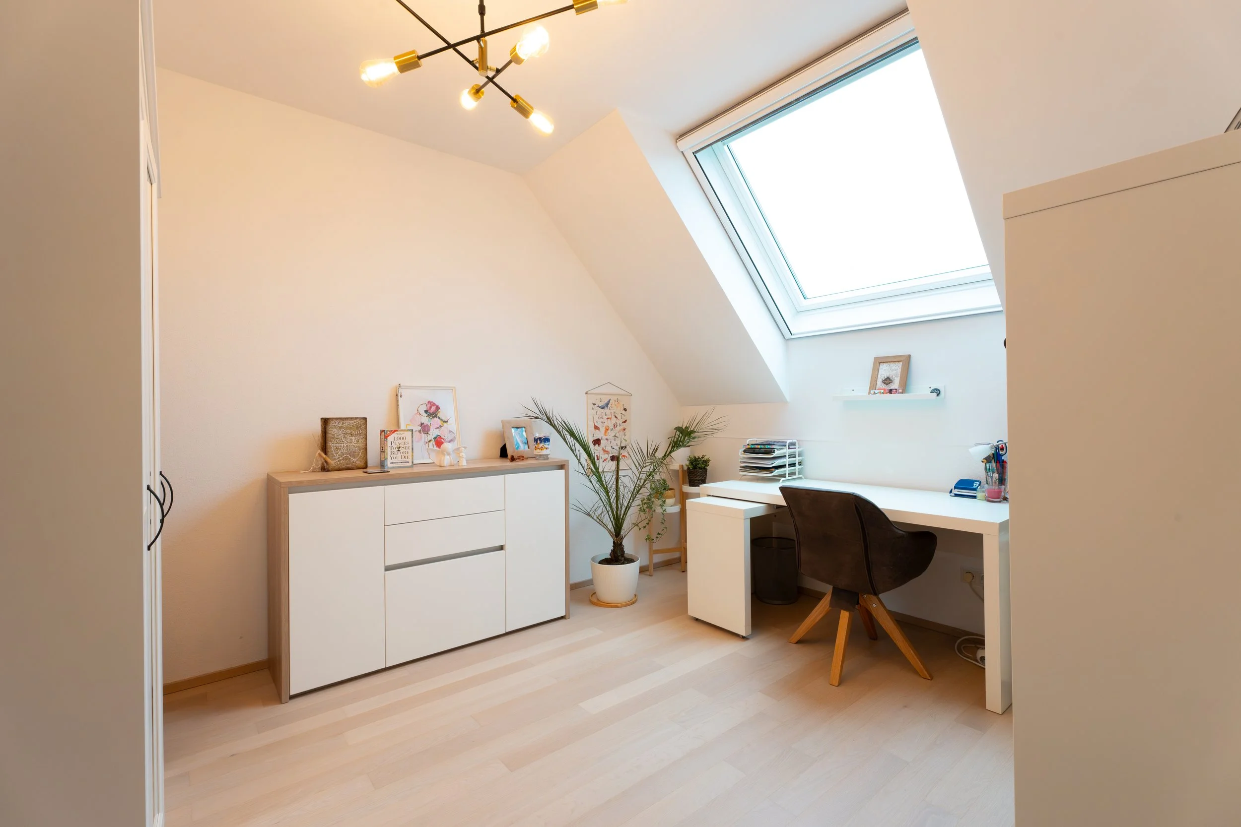 Ein helles Arbeitszimmer mit weißem Schreibtisch, schwarzem Stuhl, Dachfenster, Vasen, Pflanzen, Dekorationsbildern und einem Kleiderschrank. Die Decke hat eine moderne Leuchte.