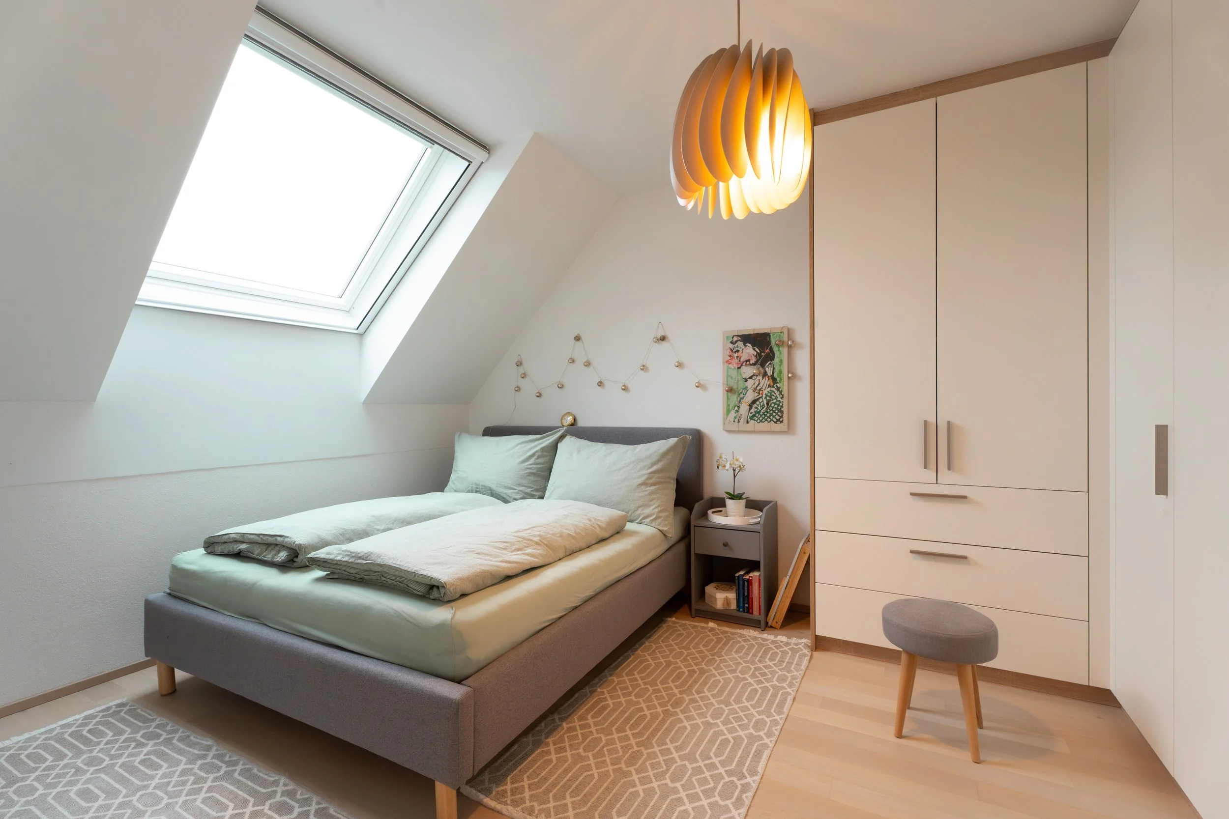 Schlafzimmer mit großem Bett, Nachttisch, Kunst an der Wand, Kleiderschrank, Teppich und Lampe, Dachfenster mit natürlichem Licht.