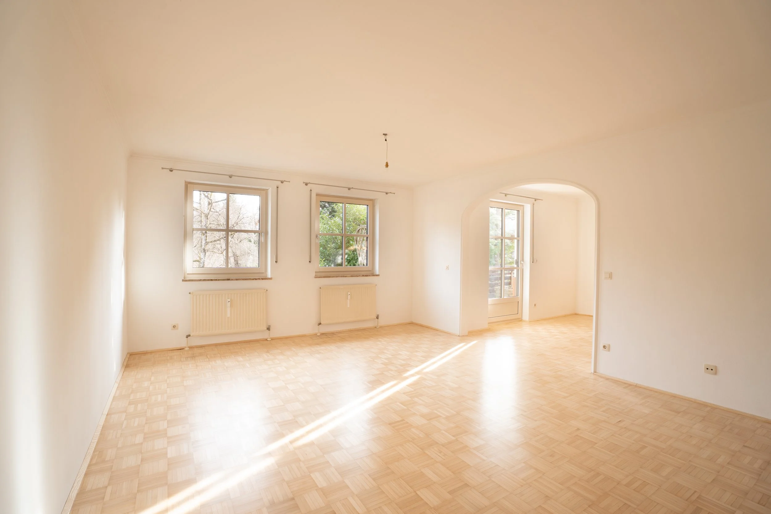 Leeres Wohnzimmer mit weißen Wänden, Holzboden, drei Fenster und einen Balkonzugang