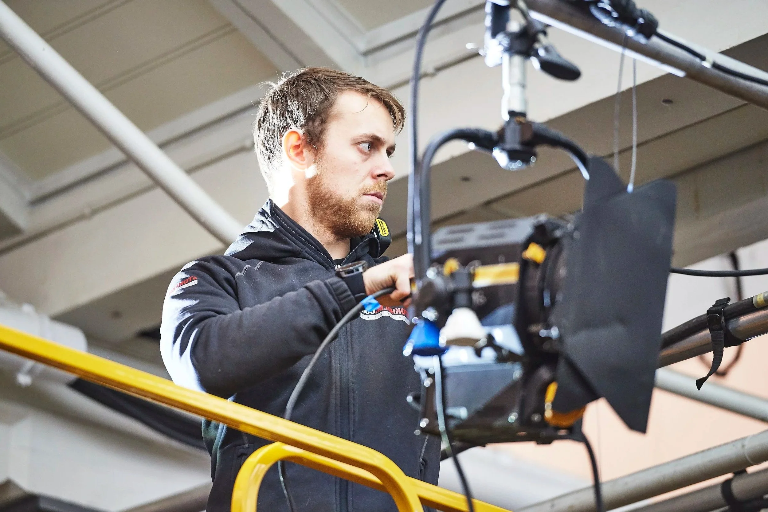A man in a black jacket with racing patches working on a piece of equipment on a lift in an industrial stage setting.