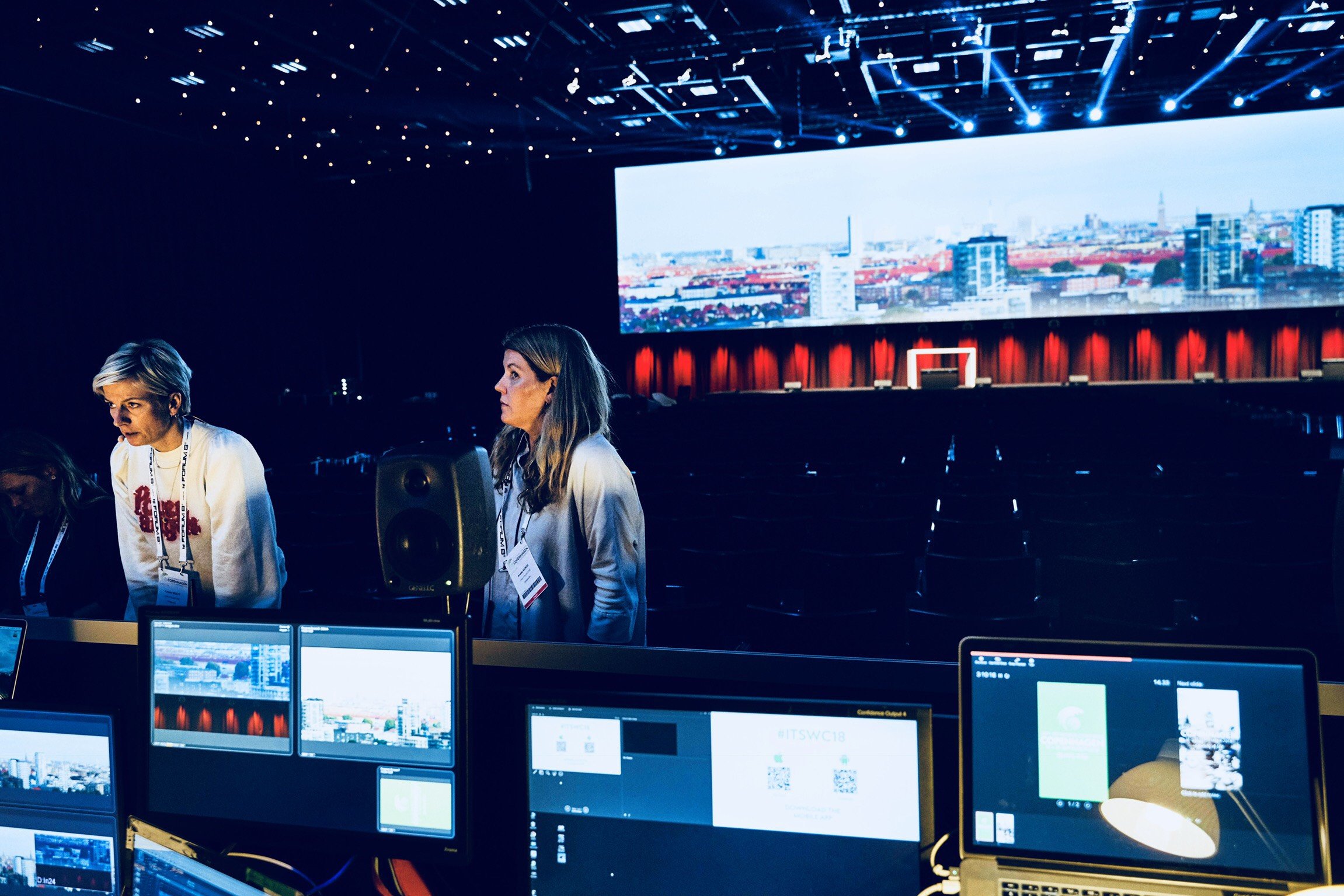 People working on computers in behind-the-scenes control room for a large event or conference, with a big screen displaying a cityscape in the background.