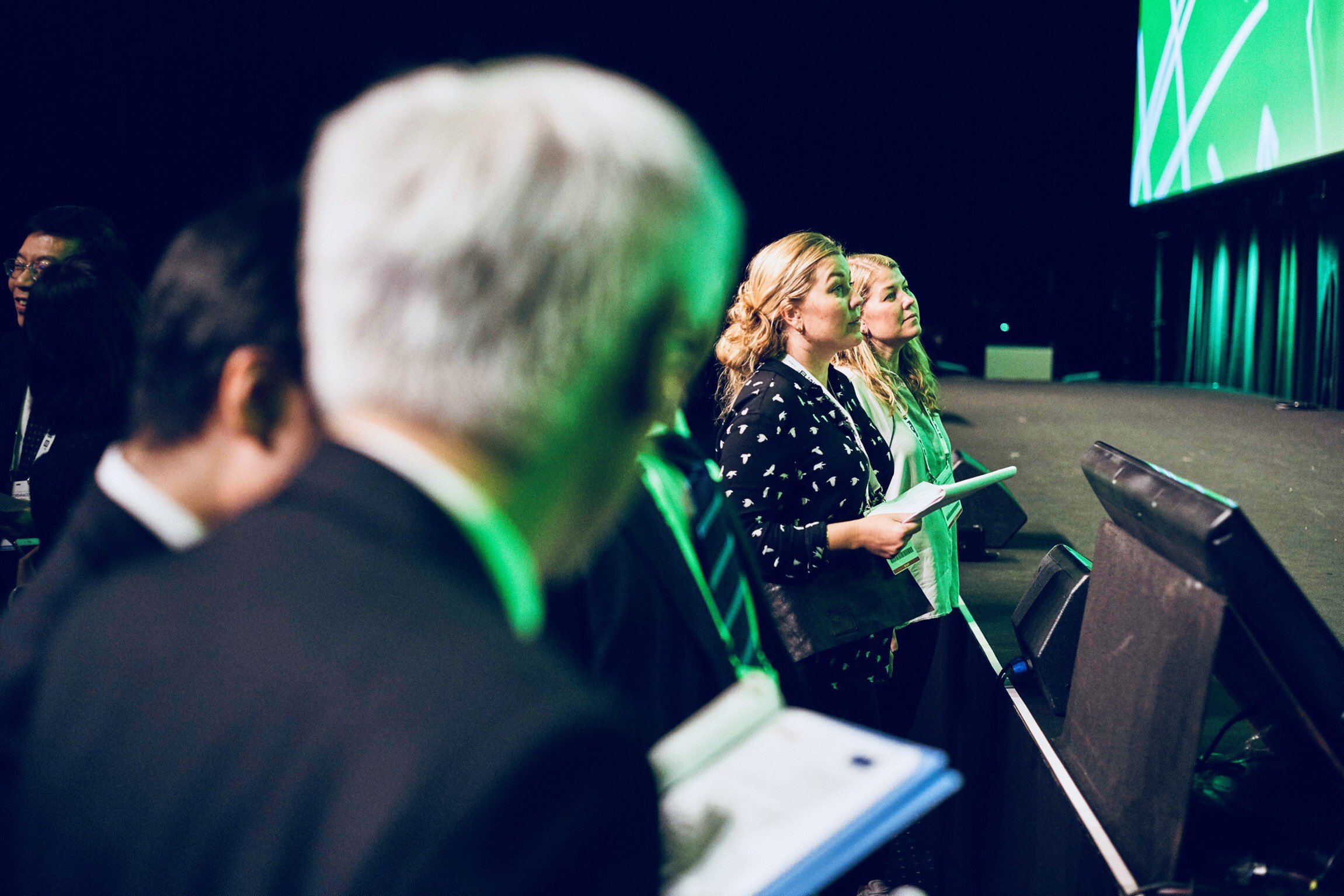 People standing in a conference or seminar, listening attentively, some holding notebooks.