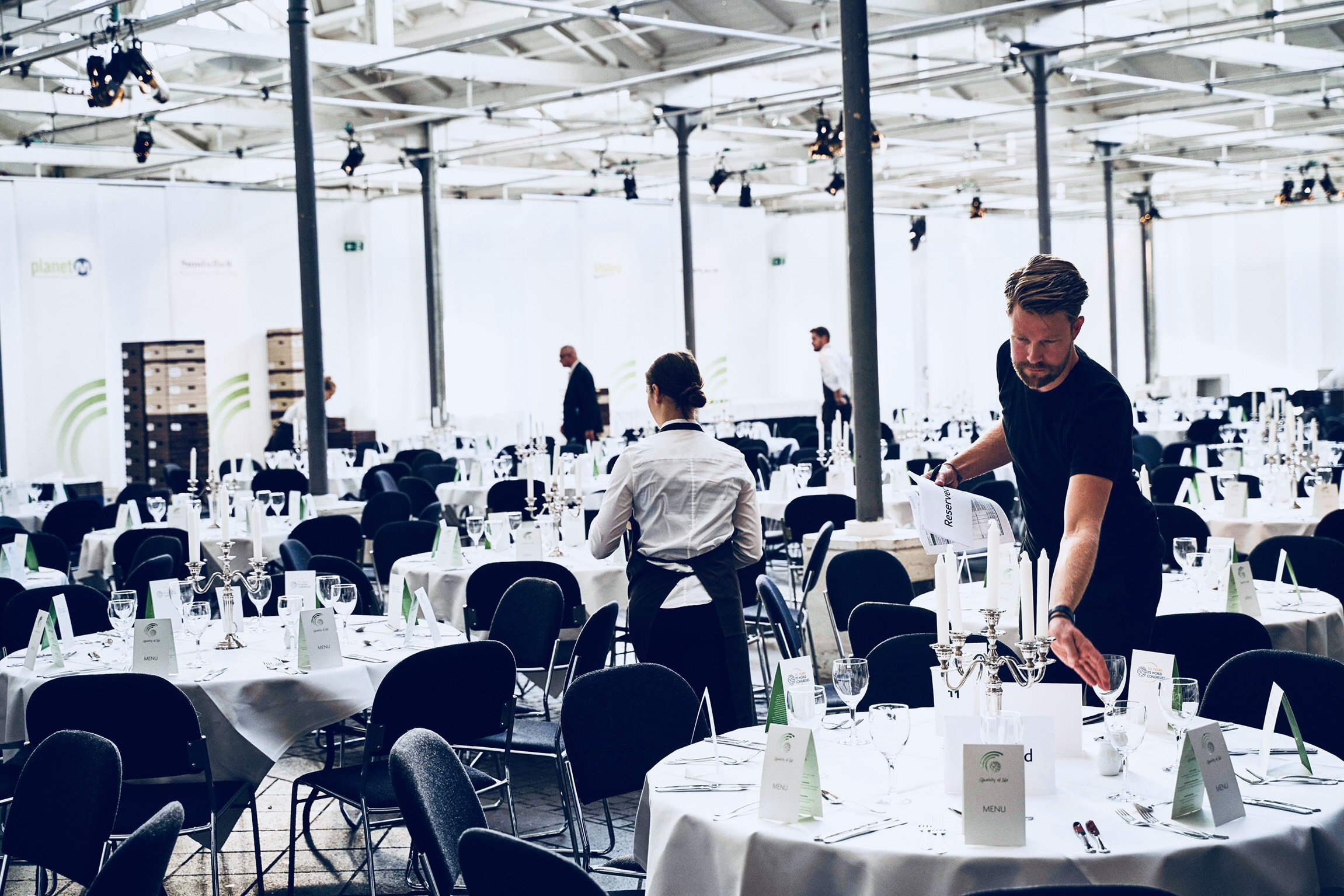 A large event venue with round tables covered in white tablecloths, set with glassware, silverware, and candles. Several waitstaff are preparing the tables, adjusting items and setting up the space.