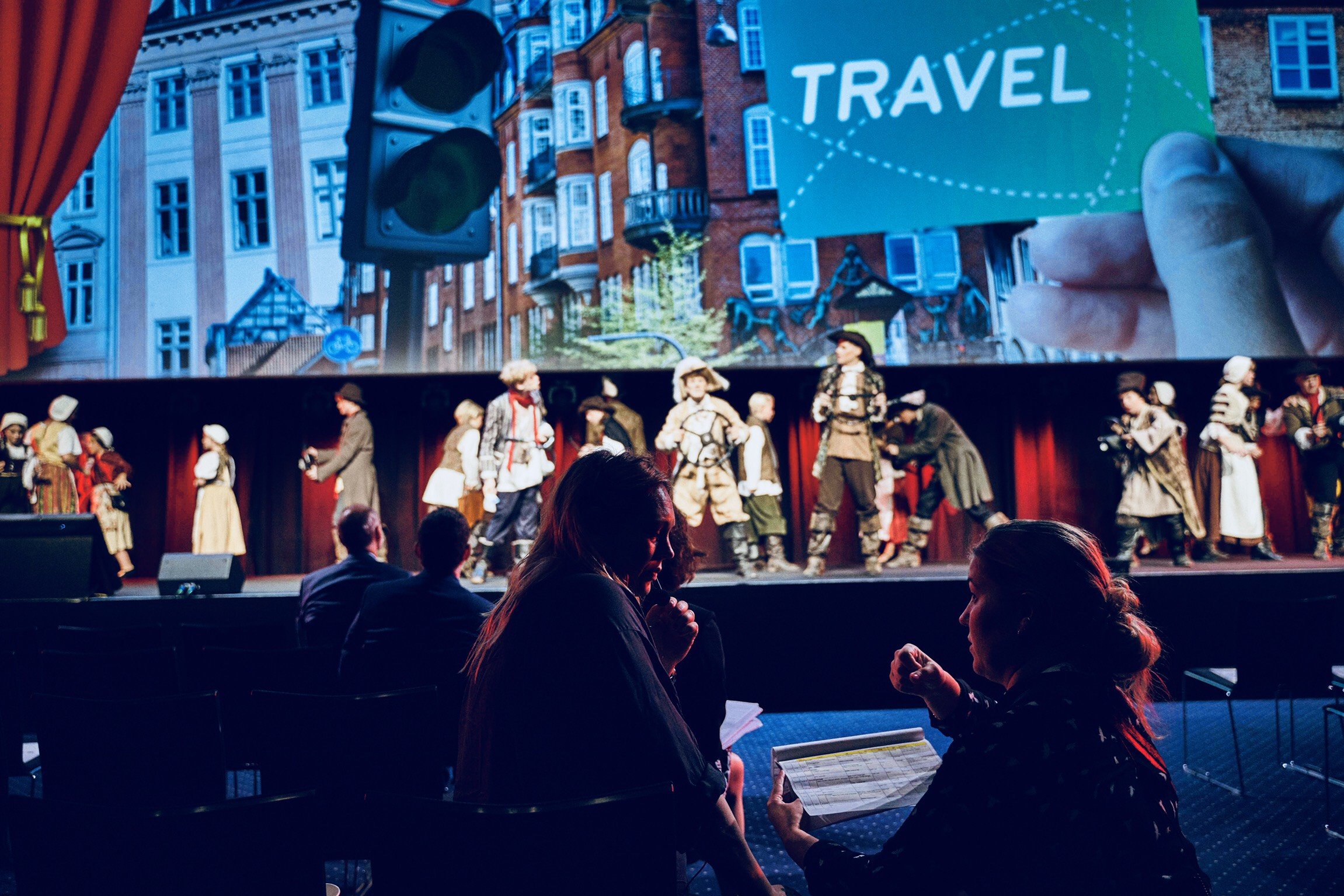 People watching a stage performance with actors in historical costumes, and a large screen projecting an image of a city street with the word "TRAVEL" and a hand holding a card.