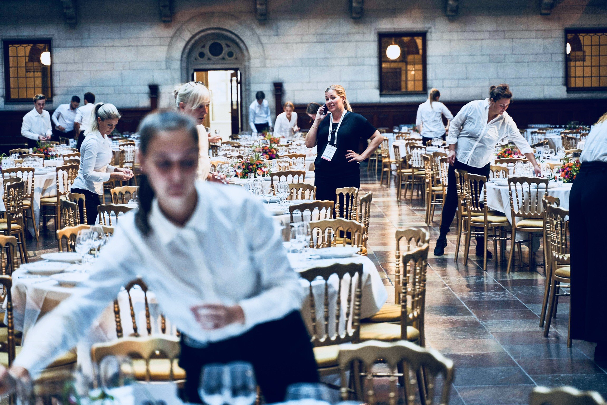 People preparing a banquet hall with tables set with white tablecloths, glassware, and centerpieces of colorful flowers.