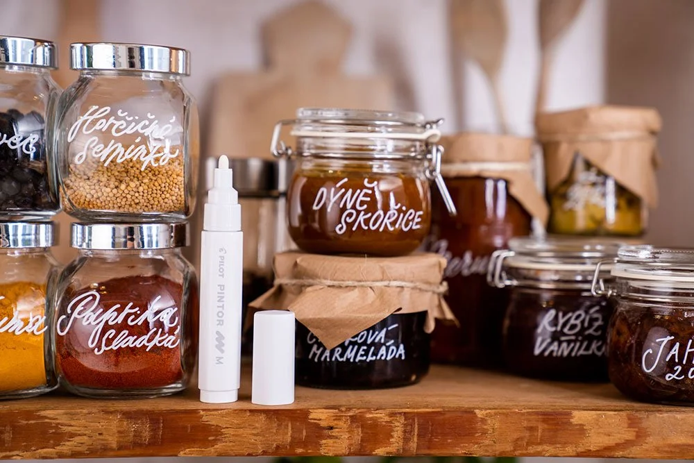 Gläser mit selbstgemachten Marmeladen, Likören und Süßigkeiten, mit Beschriftungen wie 'Hrušiče Semík', 'Jablkárská sladká', 'Olivová marmeláda', 'KYBIZ VANILKU', und 'JAHODA' auf einem Holzregal.
