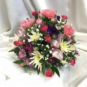 A flower centrepiece of pink, purple, yellow, and white flowers with green leaves, tied with a pink satin ribbon, resting on a white fabric surface, made by a flower shop in Crewe 'Personal Flowers'.