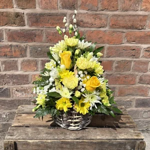 Yellow and white flower arrangement in a woven basket on a wooden surface with a brick wall background, made by a florist in Crewe 'Personal Flowers'.