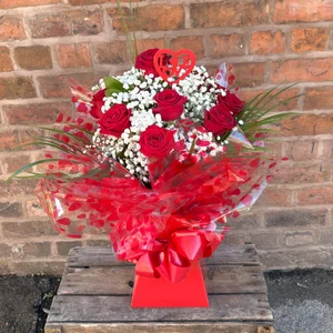 A bouquet of red roses and white baby's breath with a red heart-shaped 'LOVE' ornament on top, wrapped in clear and red heart-patterned paper, placed on a weathered wooden box against a brick wall, made by a florist in Crewe 'Personal Flowers'.