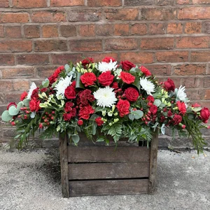 A funeral coffin spray with red roses, white chrysanthemums, red flowers and other green foliage on a wooden crate in front of a brick wall, made by a florist in Crewe 'Personal Flowers'.