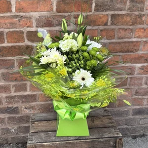 A floral arrangement with white and green flowers in a green aqua box, in decorative wrapping with green and yellow patterns, placed on a wooden crate against a brick wall background, made by a florist in Crewe 'Personal Flowers'.