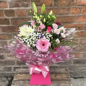A bouquet of flowers including pink roses, white lilies, white baby's breath, purple daisies, in pink decorative wrapping and tied with a pink ribbon, on a wooden box in front of a brick wall, made by a florist in Crewe 'Personal Flowers'.