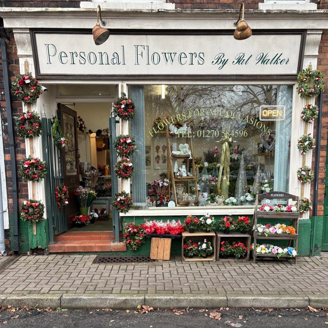 A flower shop in Crewe with a sign that reads 'Personal Flowers'. The shop is decorated with Christmas wreaths and displays various flowers and plants outside and inside. There are small crates on the right side with flower arrangements.