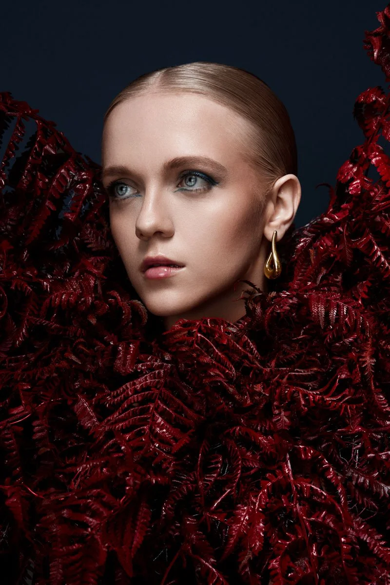 Eine junge Frau mit blauen Augen und glattem, hellbraunem Haar, die von roten Blättern umgeben ist.