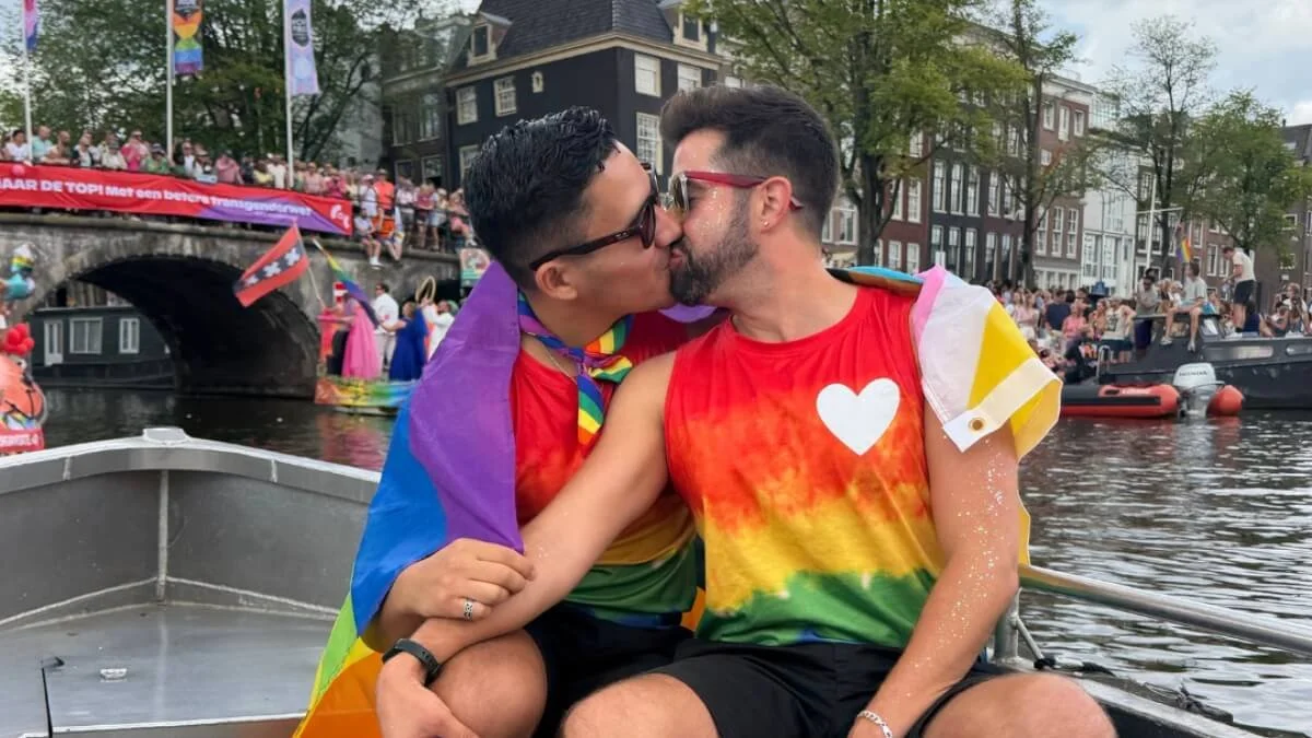 Davey and Omie kissing during Amsterdam Canal Pride with Pride crowds and boats behind them