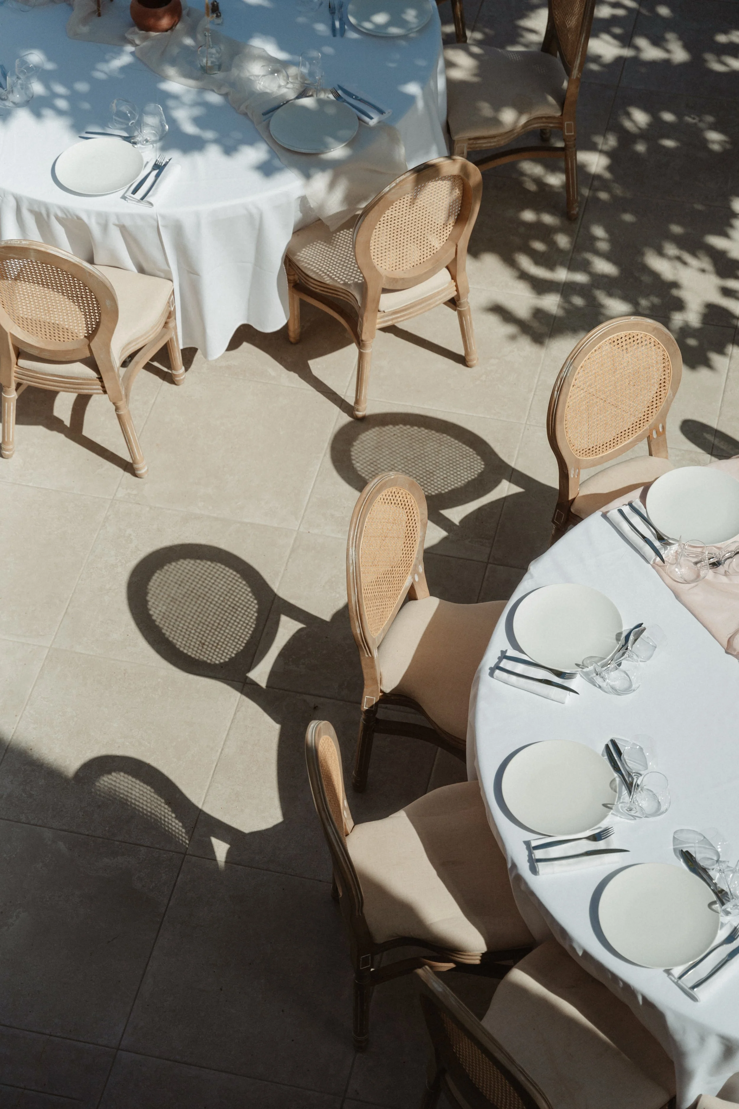 Tables de restaurant en plein air avec des nappes blanches, des assiettes, des verres et des couverts, sous des ombres d'arbres.