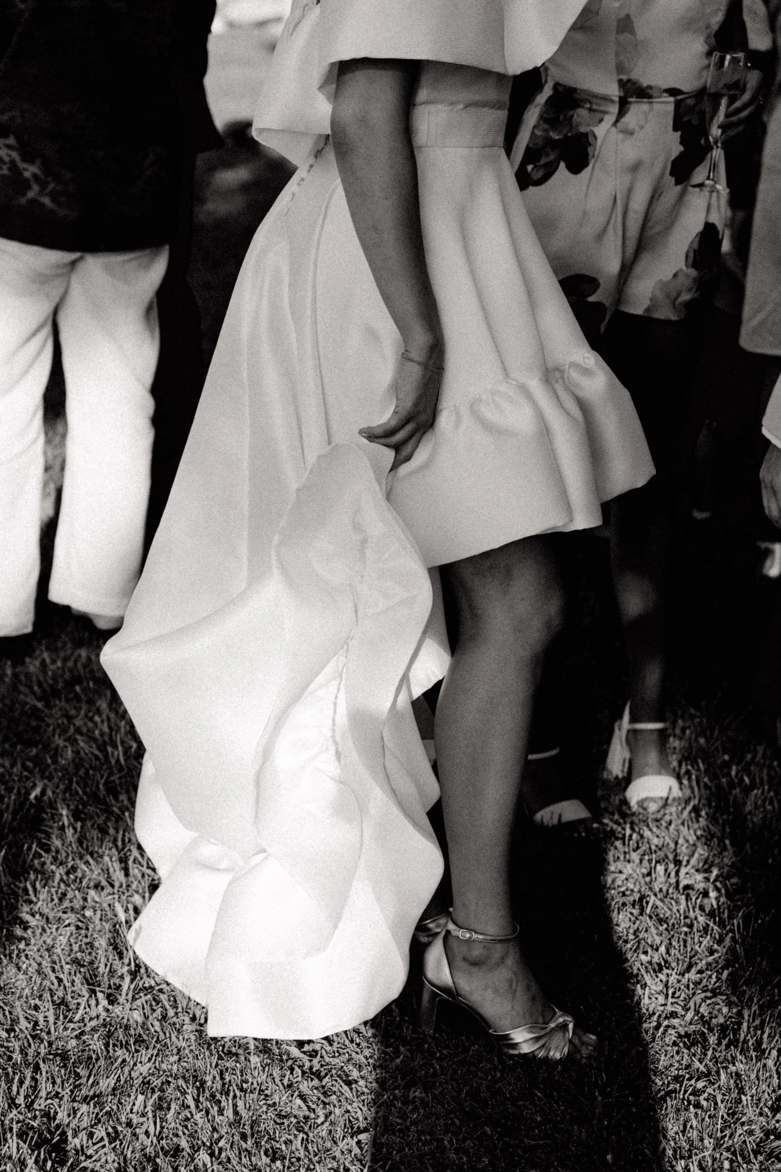 Détail d'une femme portant une robe de cérémonie, avec une jambe nue et chaussée de talons. Autres personnes en arrière-plan lors d'un événement en plein air.