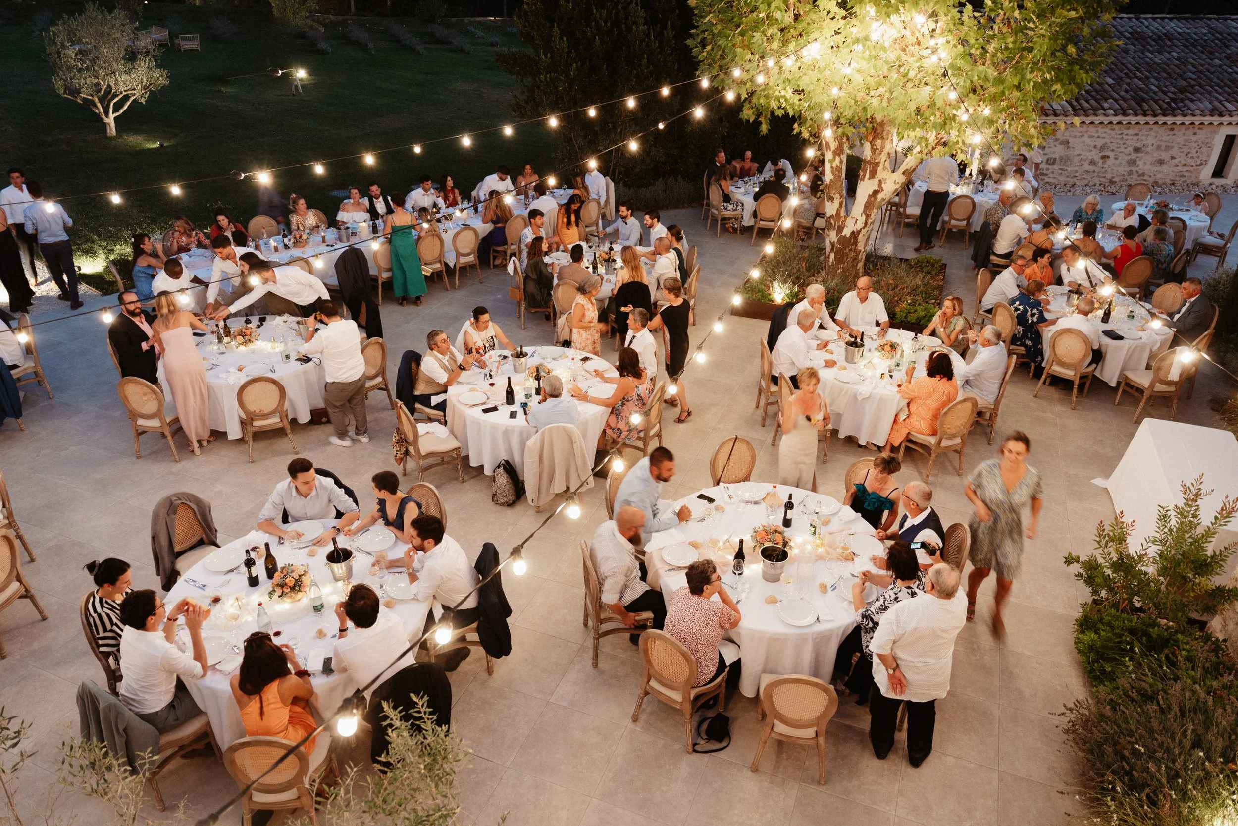 Un dîner en plein air avec plusieurs tables rondes de personnes lors d'une soirée, décorée de guirlandes lumineuses suspendues à des arbres, avec une ambiance chaleureuse et conviviale.