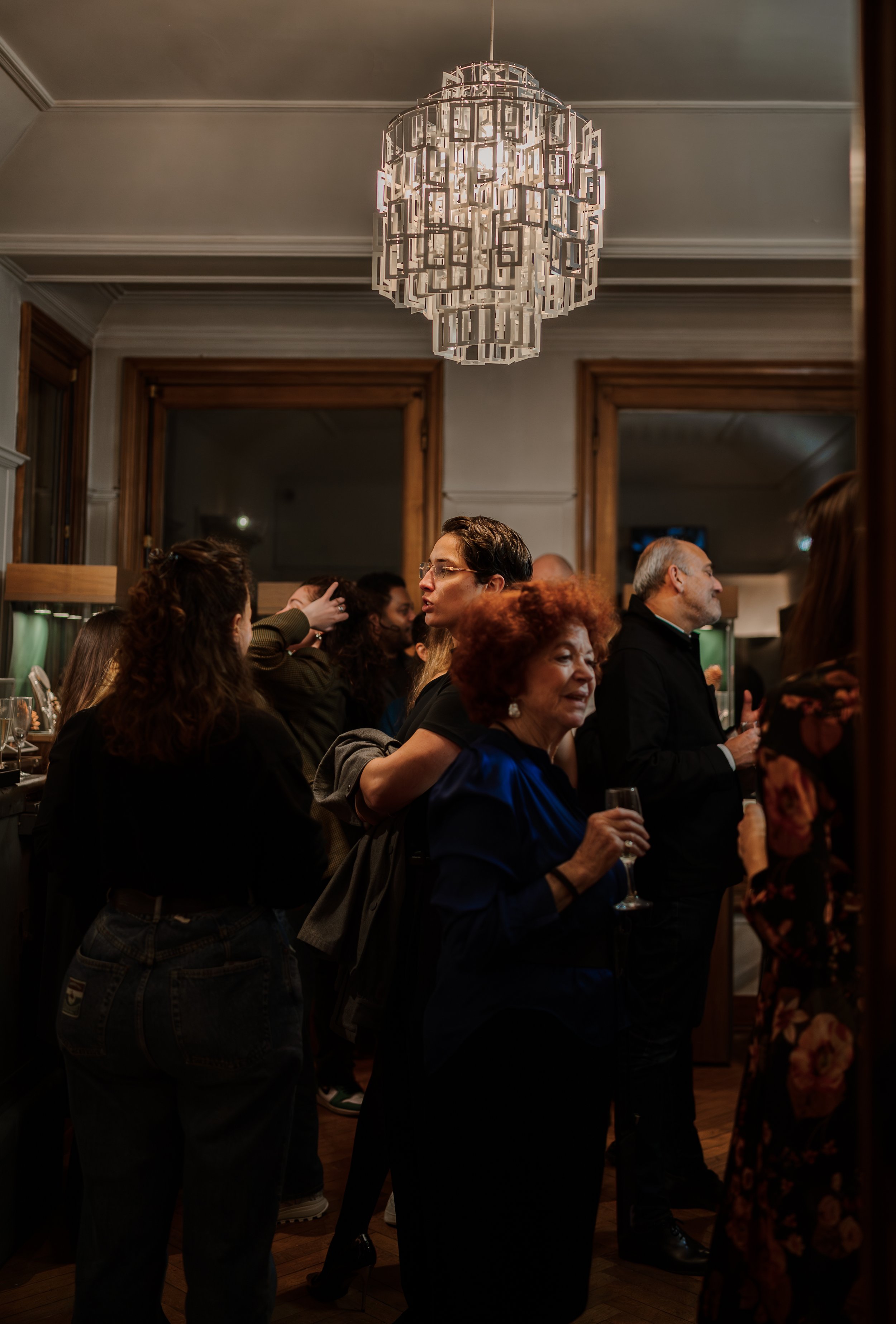 Groupe de personnes lors d'une réception ou fête dans une pièce élégante, décorée d'un lustre en métal, avec des fenêtres en bois et un plafond mouluré.