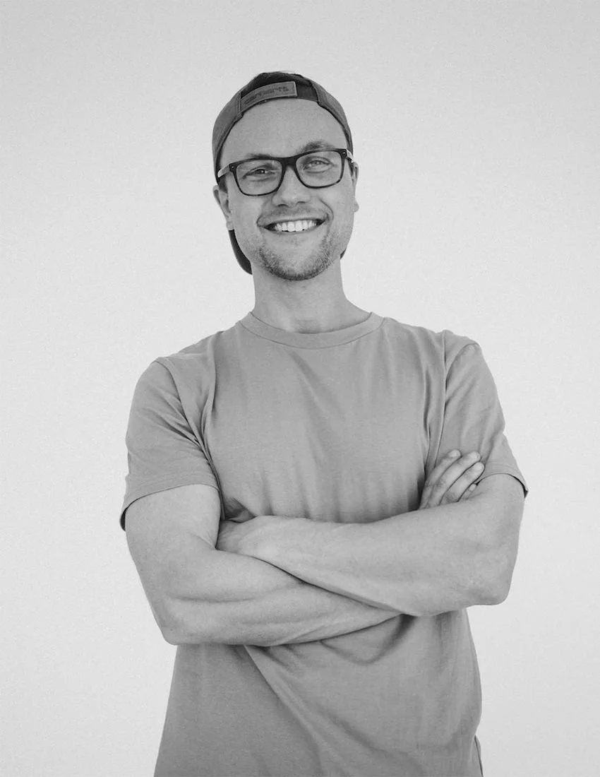 Black and white photo of a smiling person wearing glasses, a backward cap, and a T-shirt with arms crossed.
