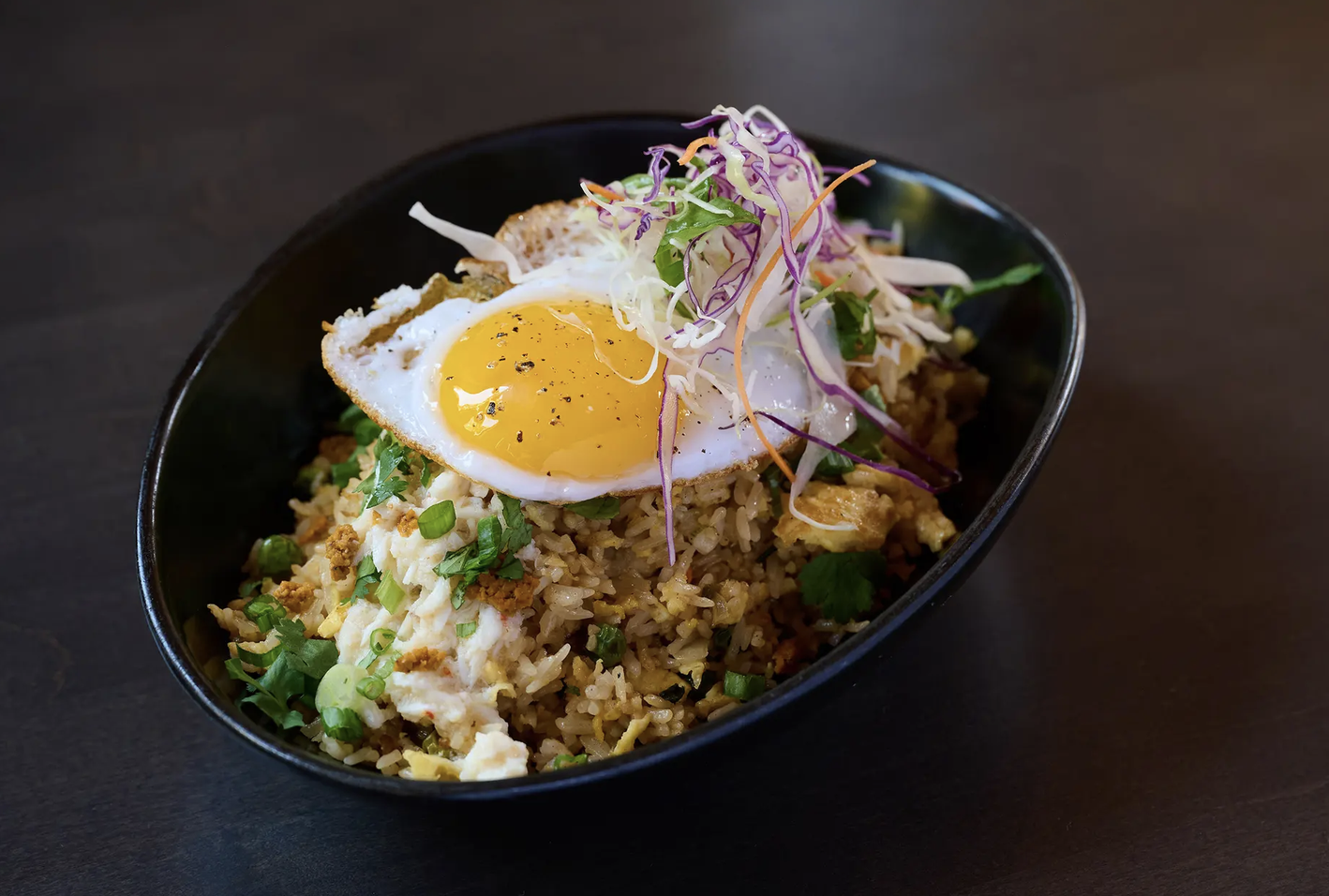 Fried rice topped with a sunny-side-up egg, fresh herbs, and shredded vegetables in a black bowl on a dark surface.