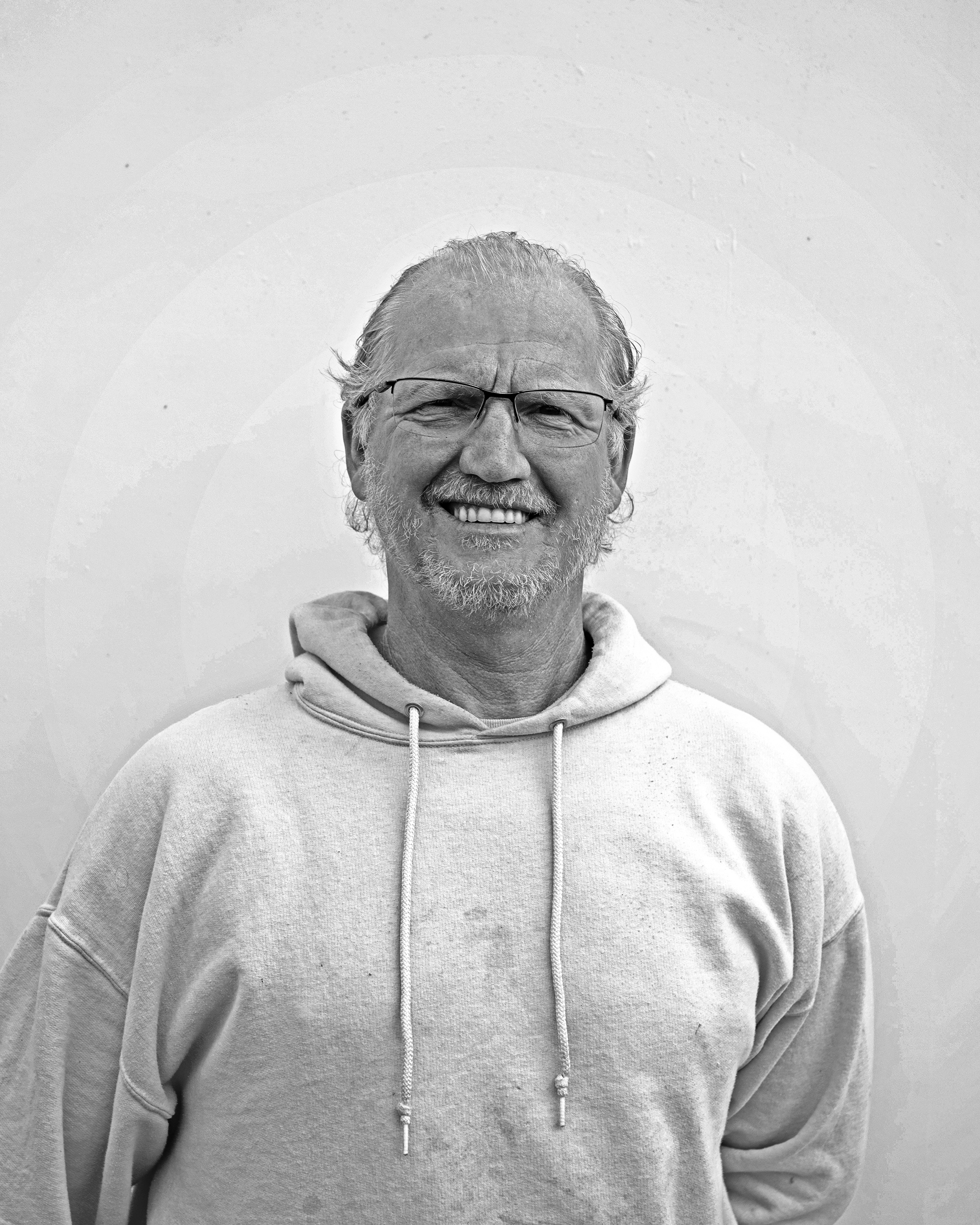 Black and white photo of a smiling man with glasses, a beard, and wavy hair, wearing a hoodie, standing against a plain wall.