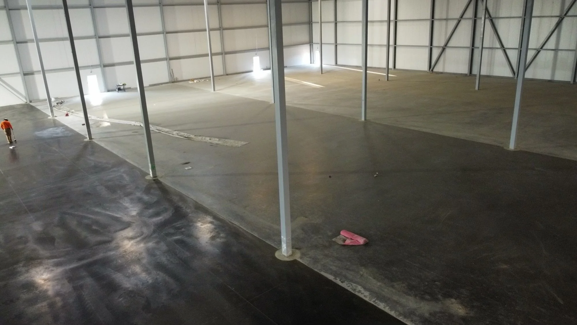 Interior of an unfinished warehouse with metal support beams, concrete floor, and construction workers in the background.