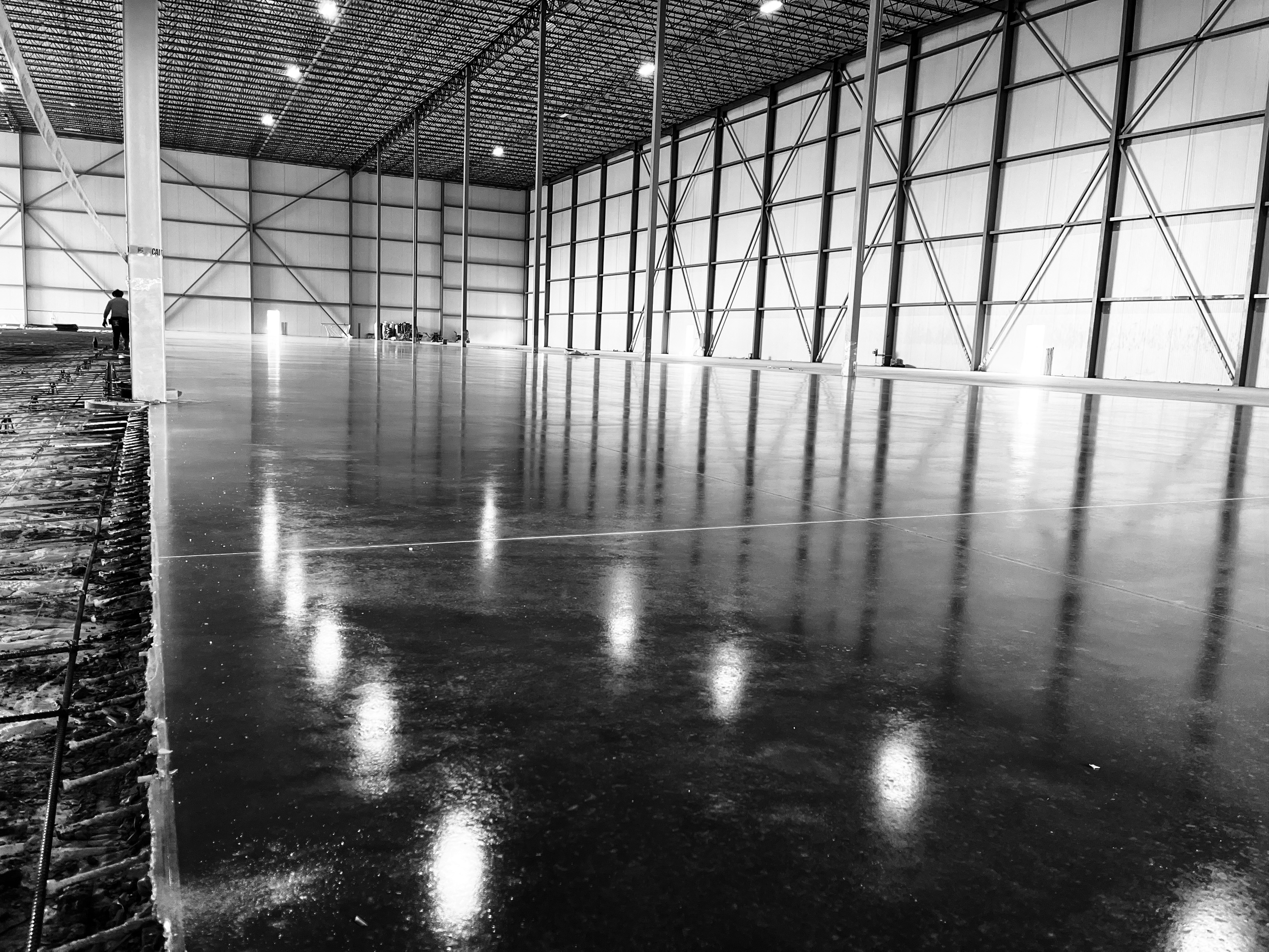 An indoor sports facility with a freshly poured concrete floor, high ceiling, and metal framework, with one person working in the background.