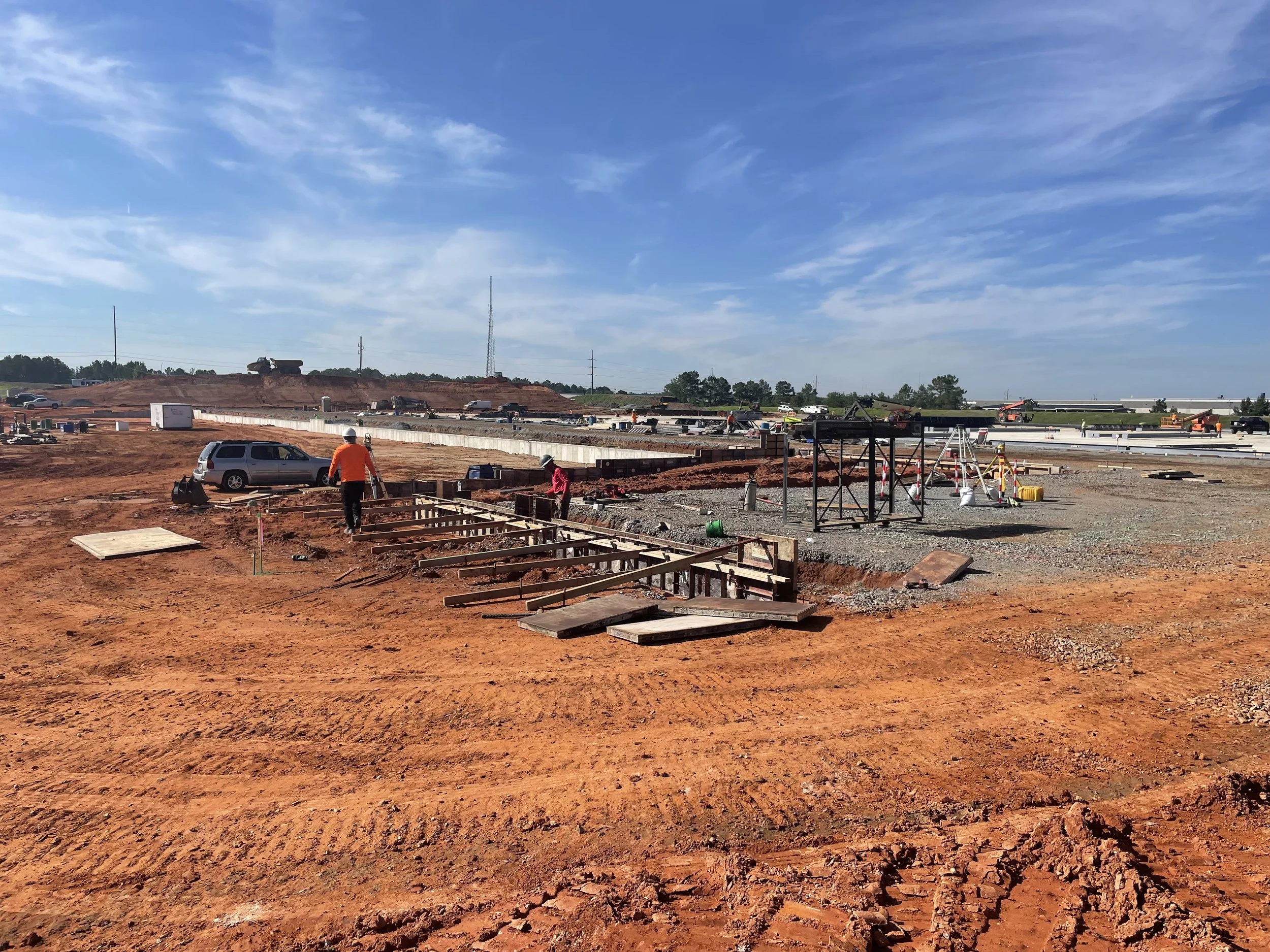 Construction site with workers, scaffolding, and construction equipment on reddish soil under a partly cloudy sky.