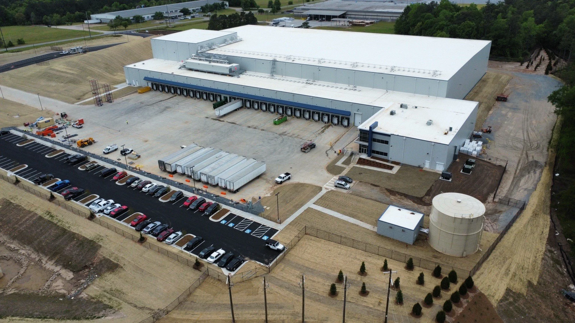 An aerial view of a large industrial building with a parking lot full of cars in front. The building has multiple loading docks and is surrounded by trees and open land.