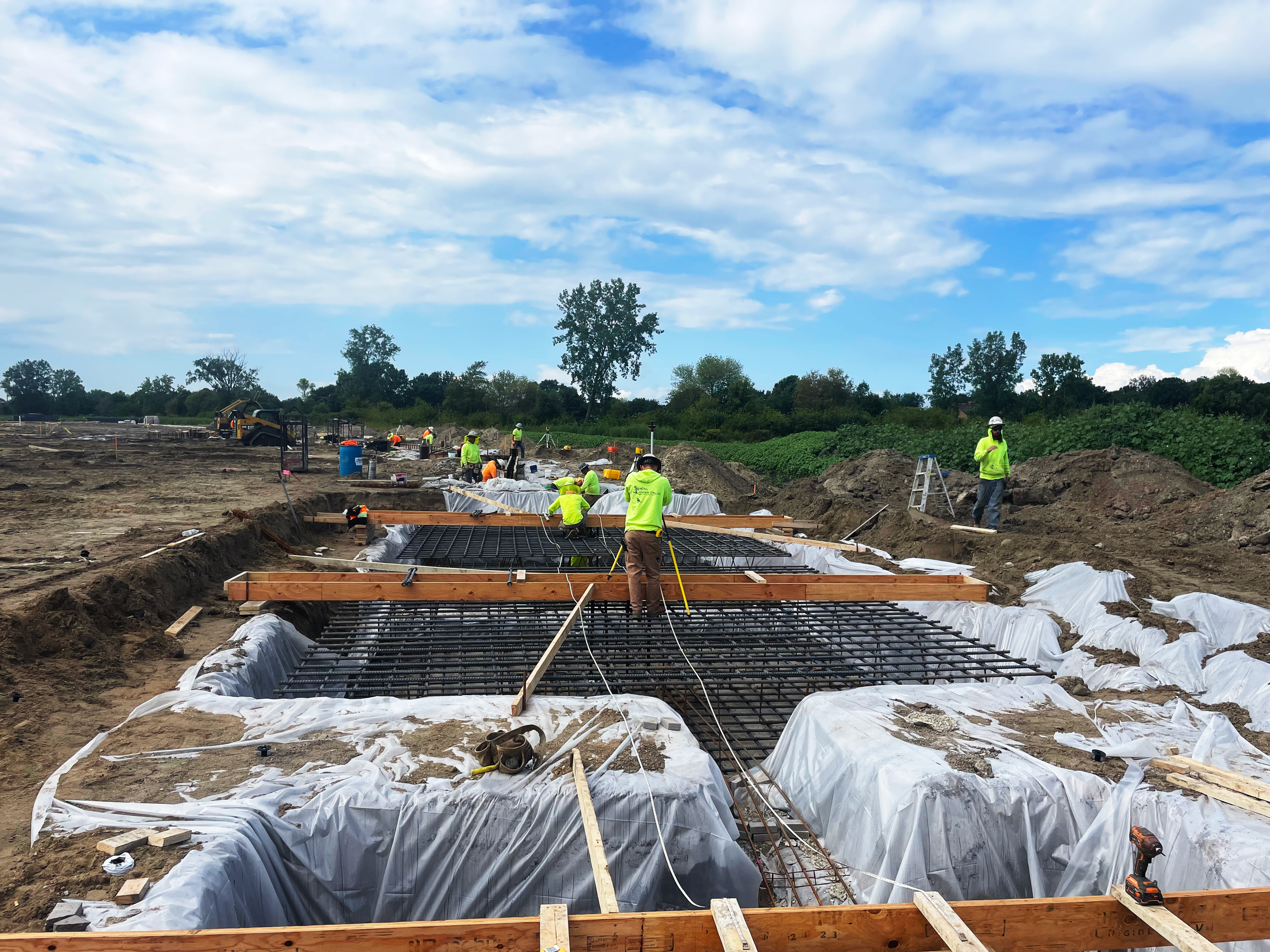 Construction workers in neon yellow shirts and hard hats working on the foundation of a building, with rebar, wooden forms, and plastic sheeting, under a partly cloudy sky.
