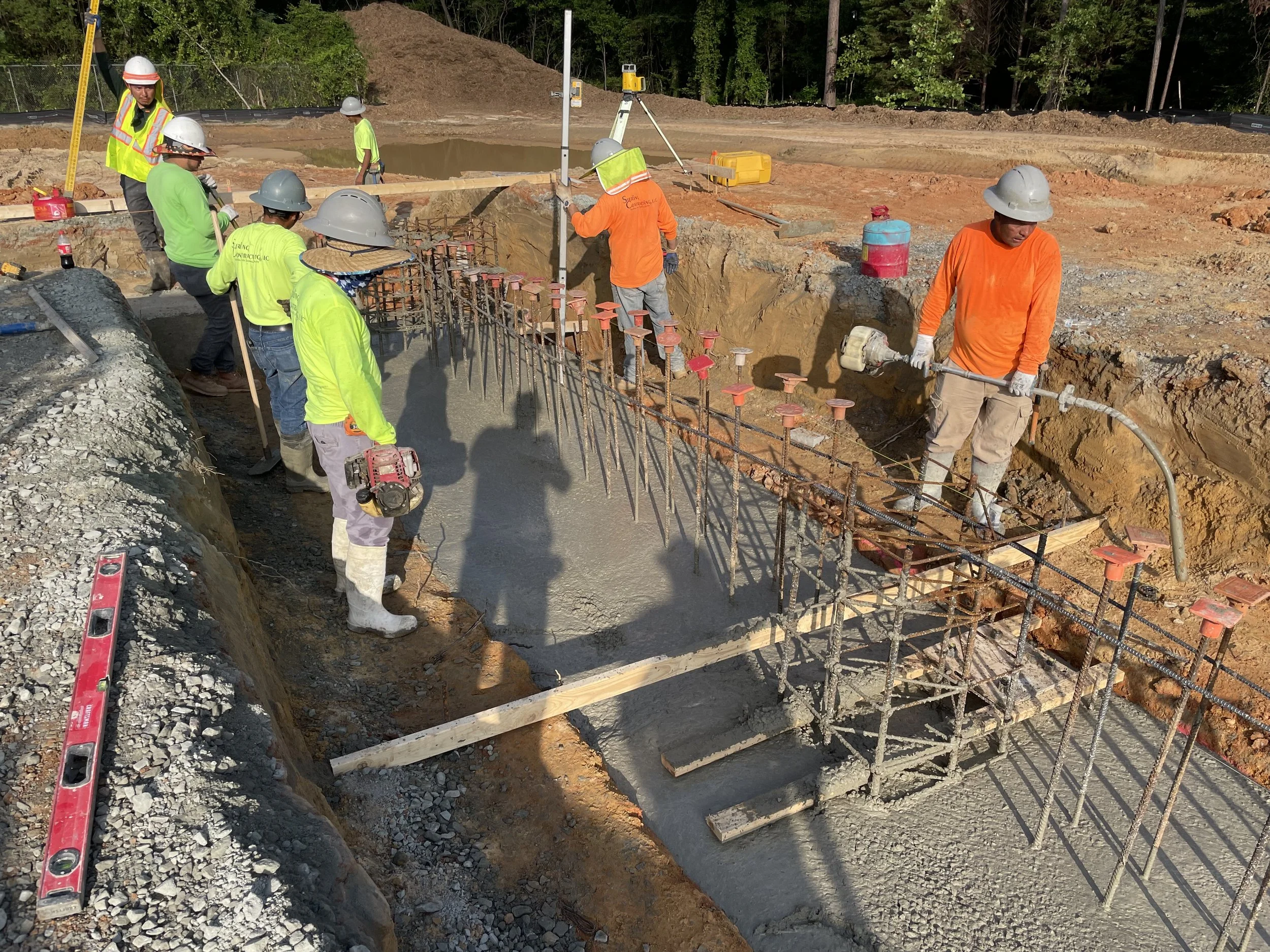 Construction workers wearing safety helmets and high-visibility clothing pouring concrete into a foundation with rebar reinforcement at a construction site surrounded by trees.