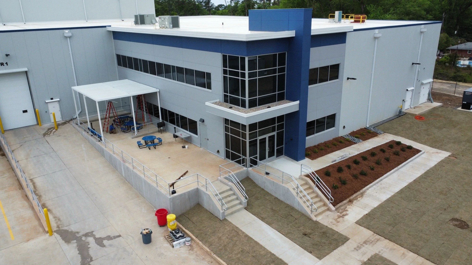 A modern industrial building with a blue and light gray exterior, featuring a large entrance with glass panels and an outdoor play area with swings, tables, and benches. There are landscaping and newly laid grass patches, with some construction equip