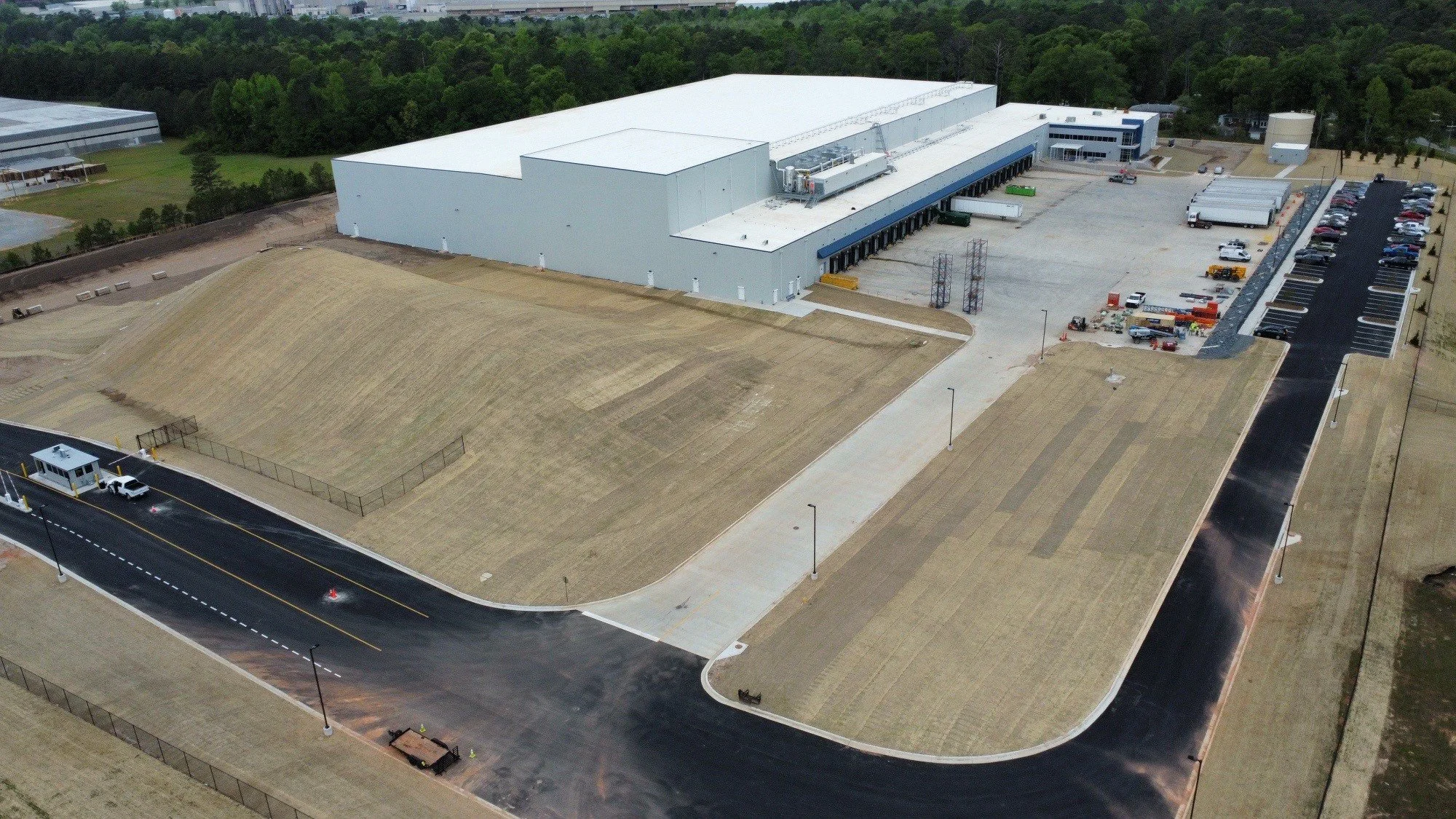 An aerial view of a large industrial or warehouse building with a white roof, surrounded by parking lots and roadways, and some grassy areas and trees in the background.