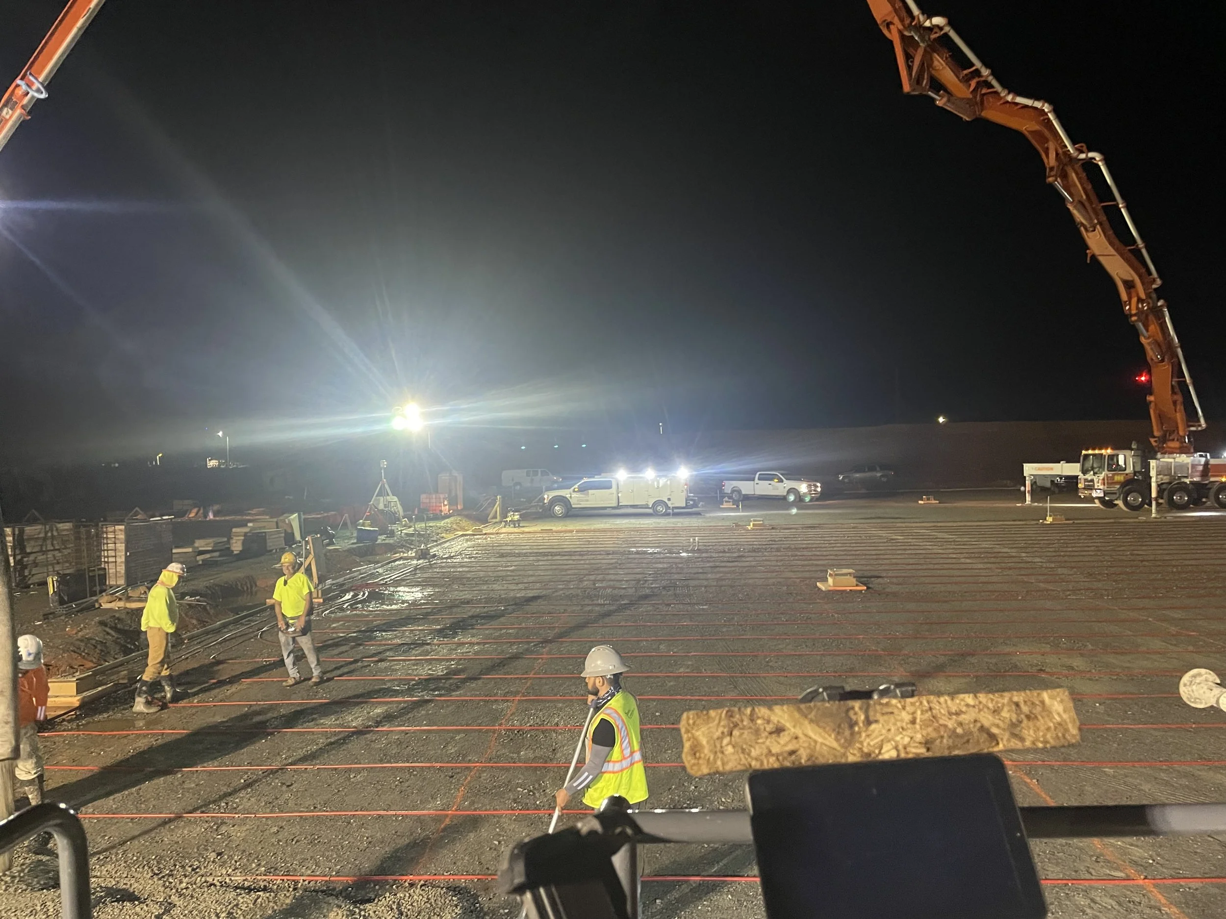 Nighttime construction site with workers in safety vests and helmets, concrete being poured or prepared, construction trucks, and large equipment under bright lights.