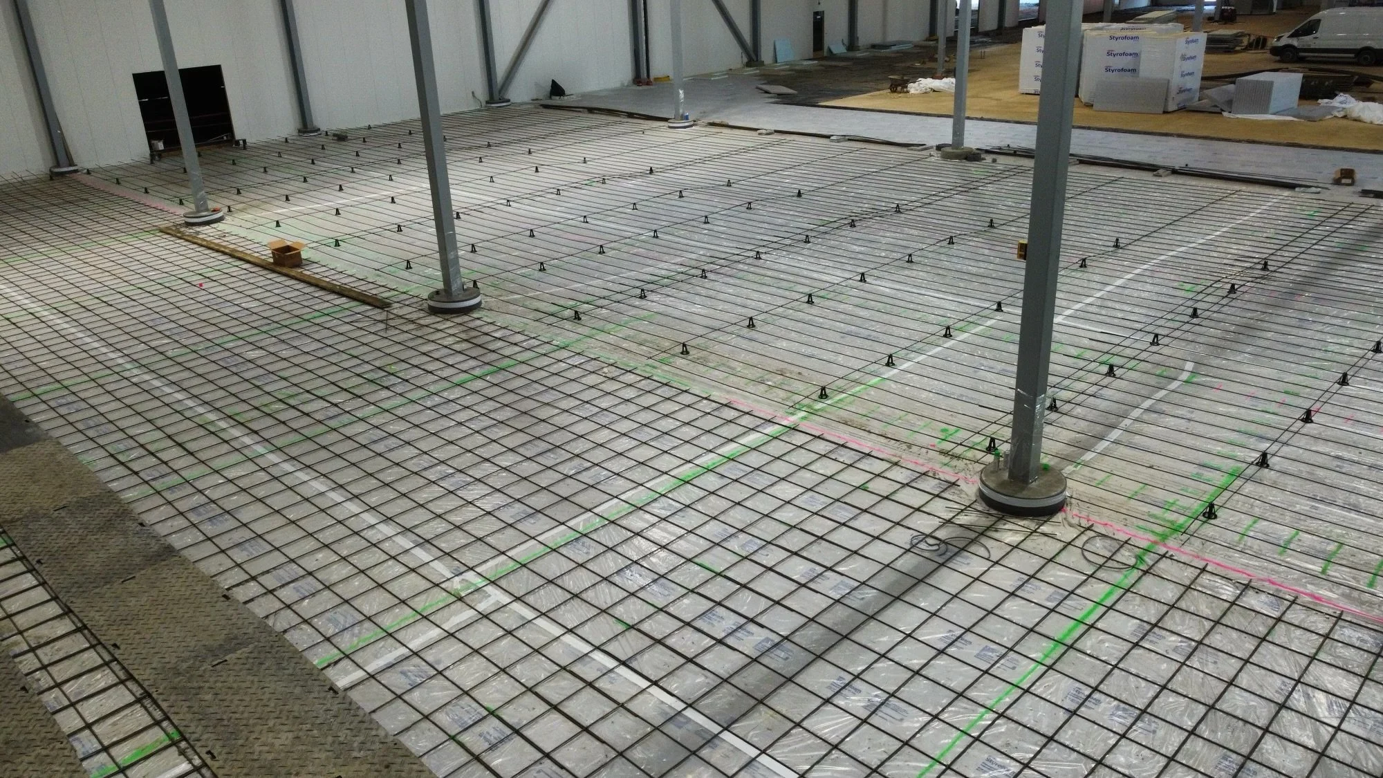 Construction site with steel rebar grid for concrete pouring, supporting columns, and building materials in the background.