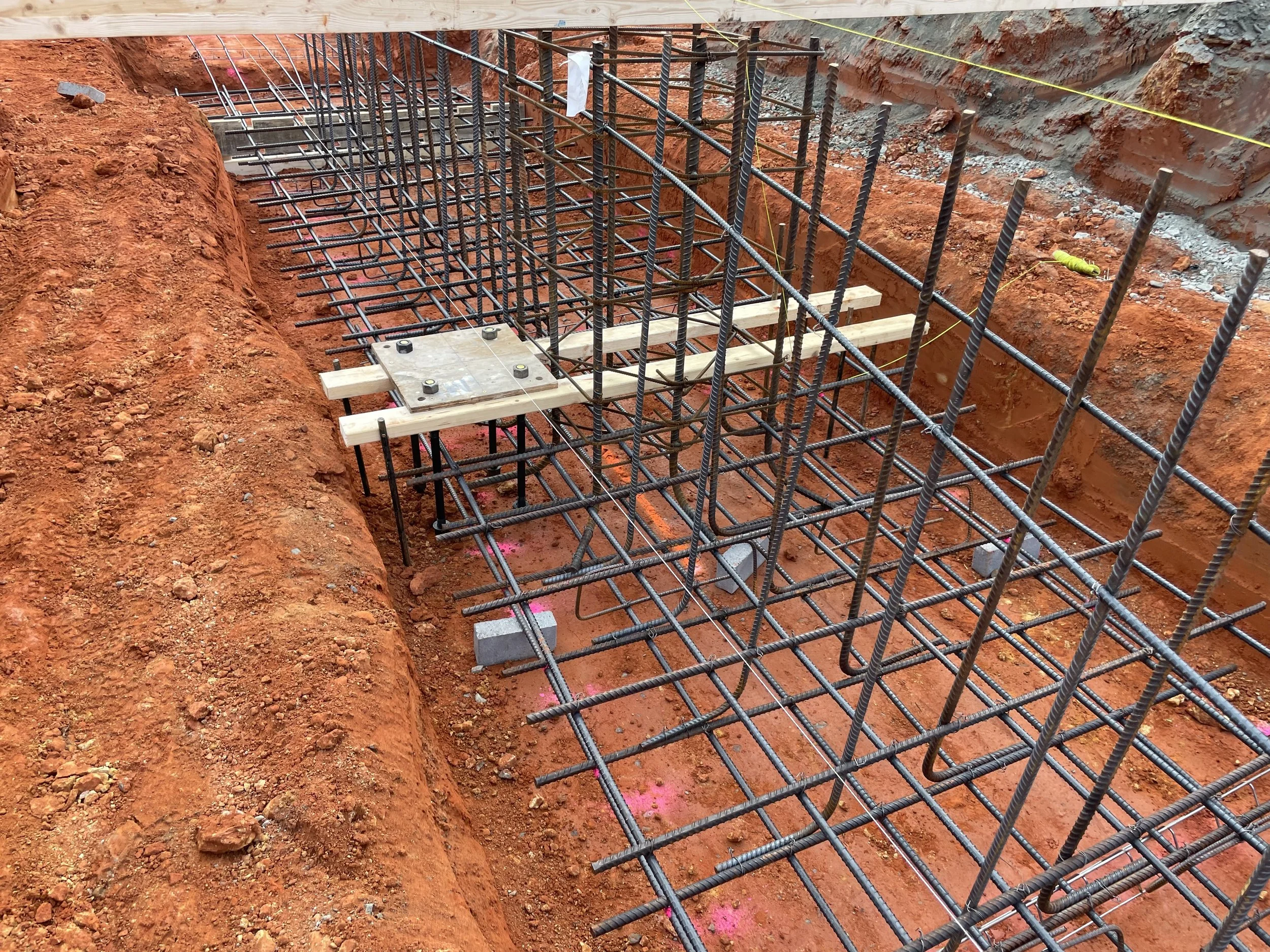 Construction site with exposed rebar and wooden supports for a foundation, showing a deep excavated trench with orange soil.