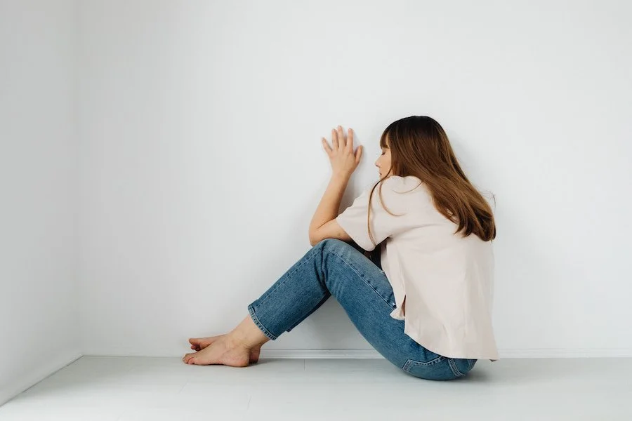 A contemplative woman sitting barefoot against a white wall, conveying solitude.